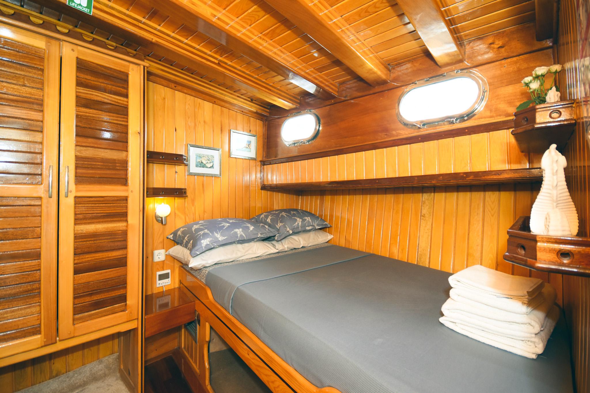 Gulet double cabin with varnished wood panelling, double bed with blue pillows, louvred wardrobe, and oval porthole windows
