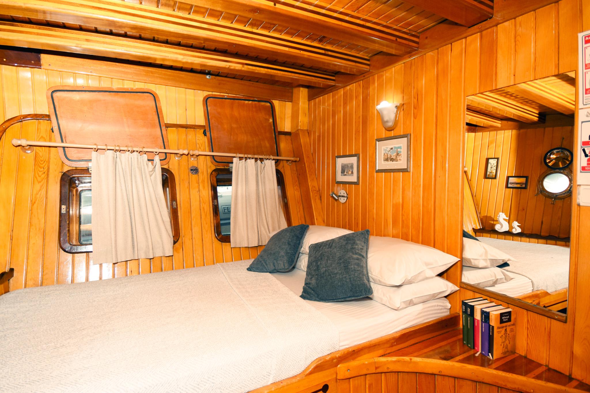 Gulet double cabin with varnished wood panelling, two portholes with curtains, white bedding and blue cushions