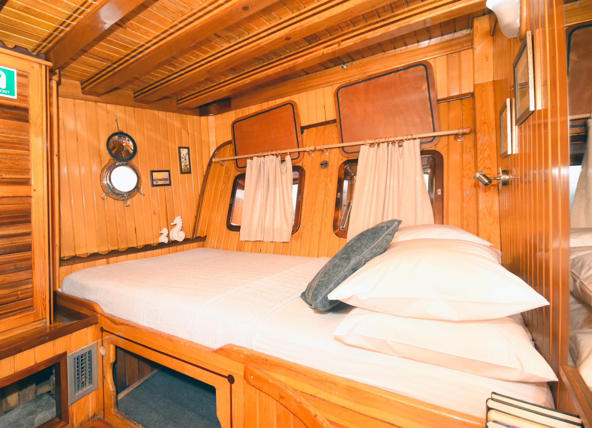 Gulet double cabin with warm wood panelling, porthole, two opening hatches, and white bedding with grey accent pillow