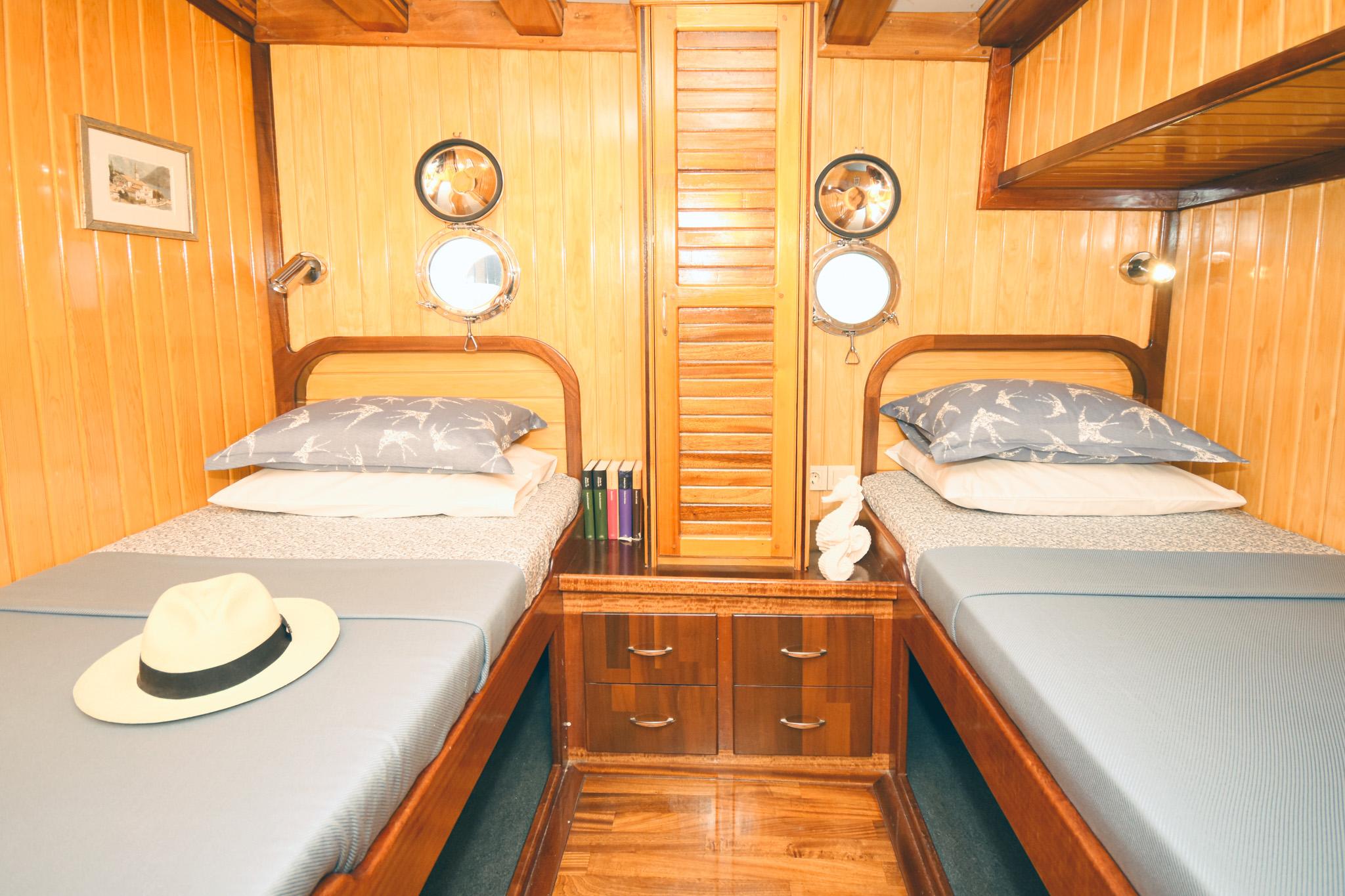 Gulet twin cabin with two single beds, matching grey pillows, polished teak panelling, dual portholes, and central louvred wardrobe door