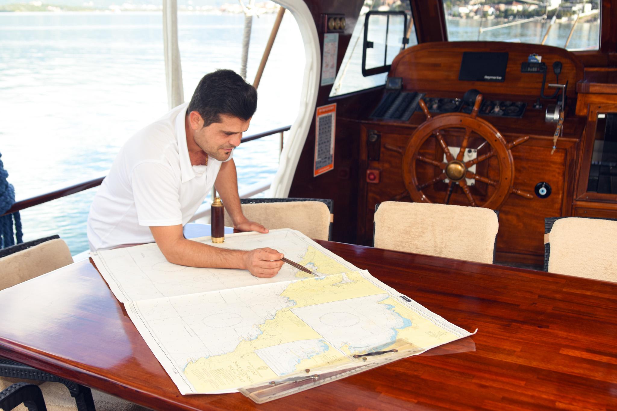 Gulet captain studying nautical chart at dining table beside traditional wooden helm station