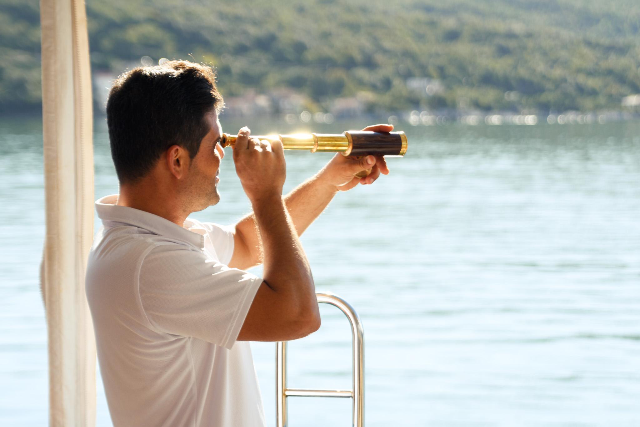 crew member in white polo shirt looking through brass handheld telescope from yacht deck with green hillside coastline behind