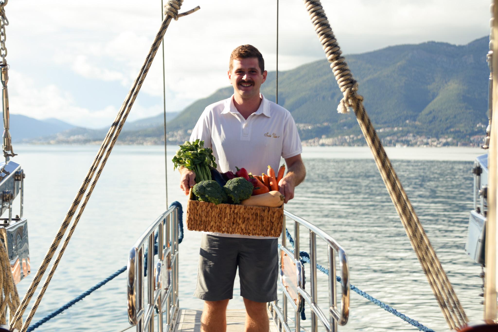 smiling crew member holding wicker basket of fresh vegetables on gulet stern boarding platform with mountain backdrop