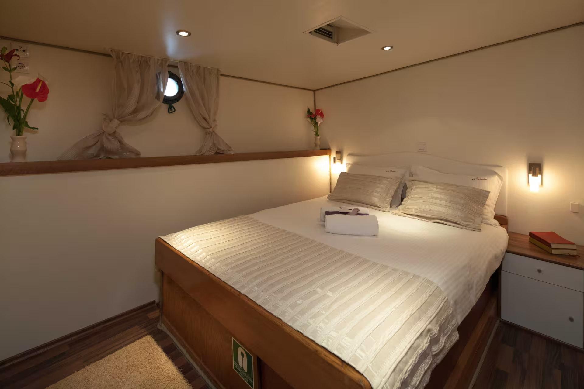 Gulet double cabin with wood-framed bed, white bedding, wall-mounted reading lights, porthole window with tied curtains and fresh flowers