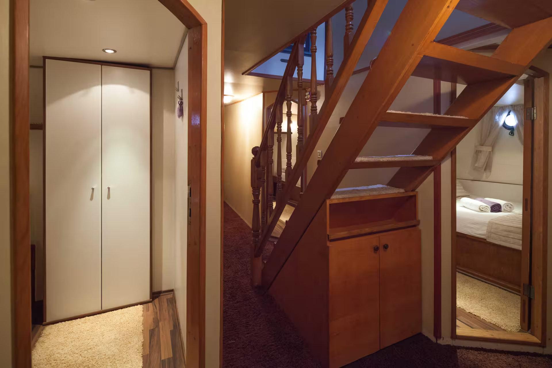 Gulet below-deck corridor with wooden staircase, built-in storage cabinet, and open cabin doorway revealing a made bed with folded towels