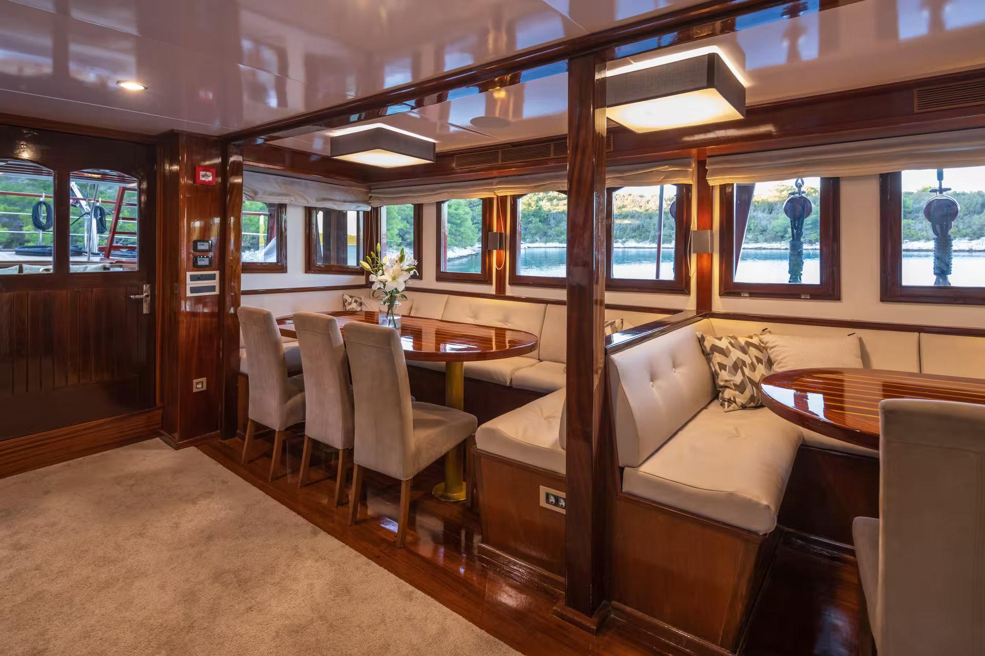 Gulet main saloon with polished wood oval dining table, upholstered chairs, and cream tufted banquette seating with panoramic side windows