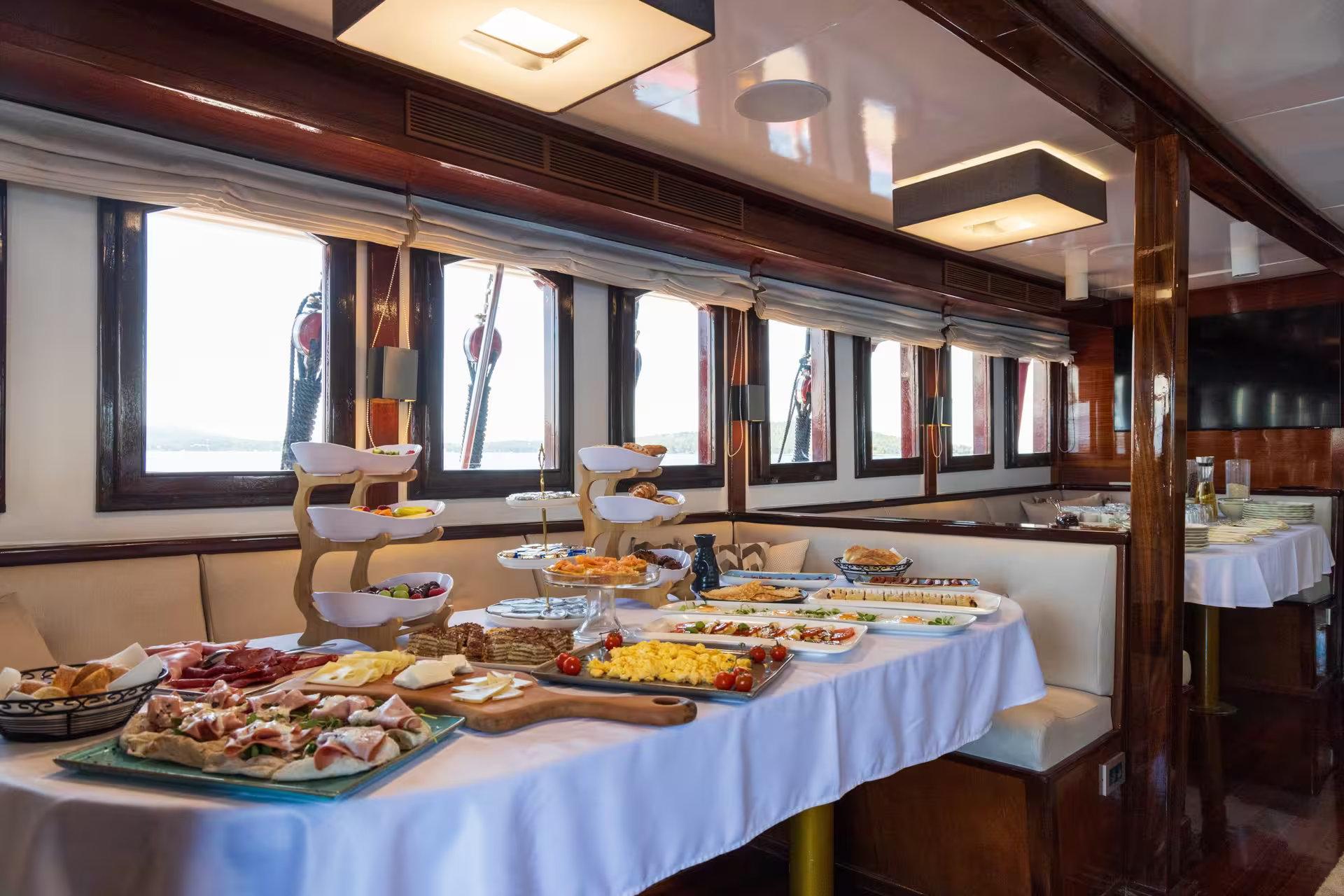 Luxury gulet indoor dining salon with full breakfast buffet spread including charcuterie, cheeses, pastries and tiered fruit stands