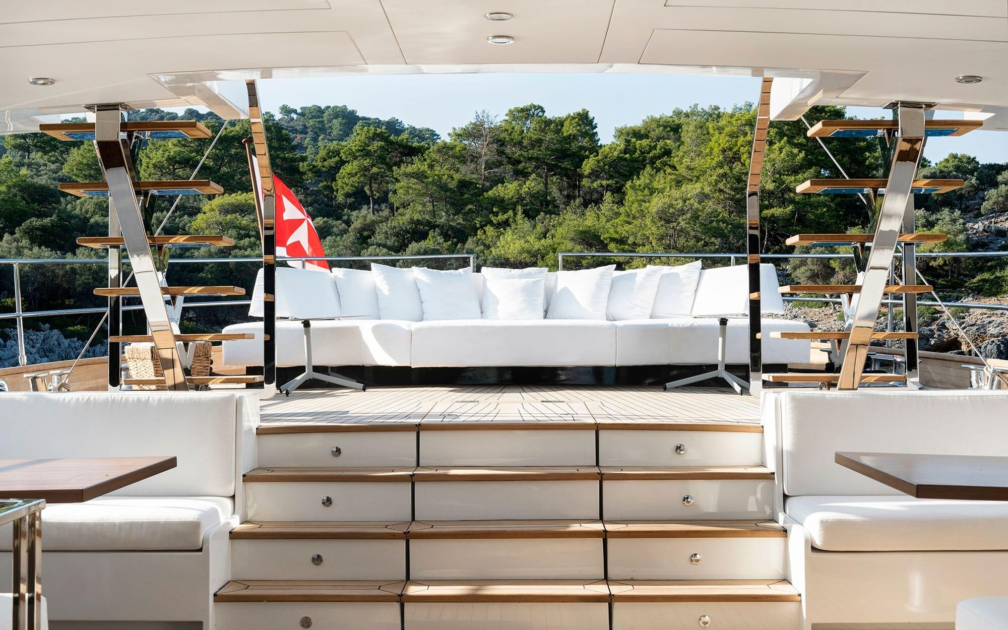 yacht upper deck with white outdoor sofa, teak stairs, and forest coastline view