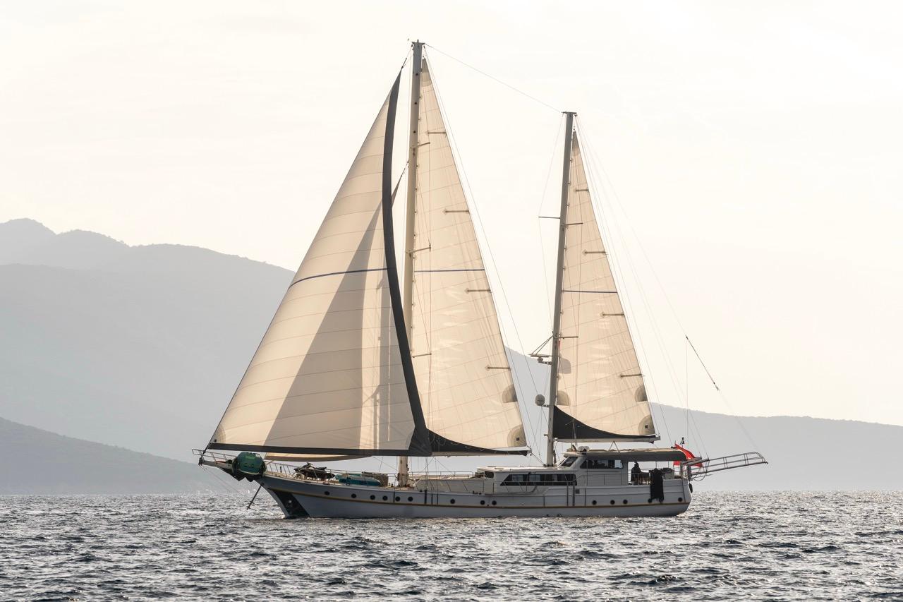 white sailing gulet with full cream sails deployed cruising coastal waters near mountainous coastline