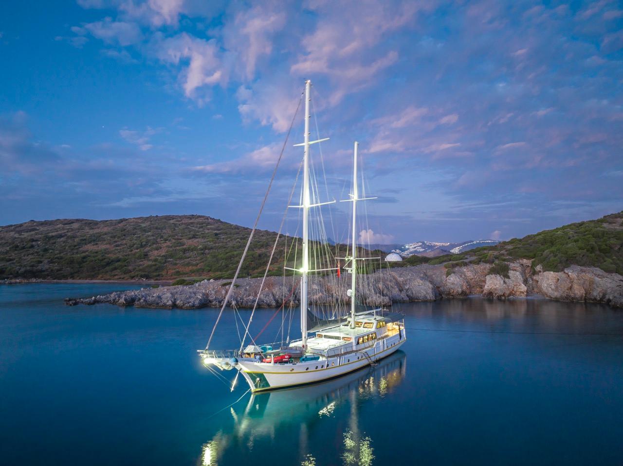 White traditional gulet with two masts anchored in calm blue bay surrounded by rocky coastline at dusk