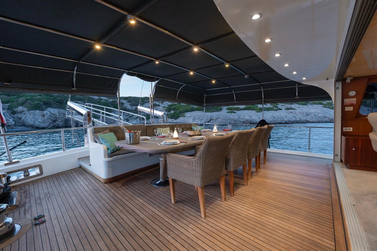 Covered aft deck with dining table, wicker chairs and built-in seating overlooking coastal waters