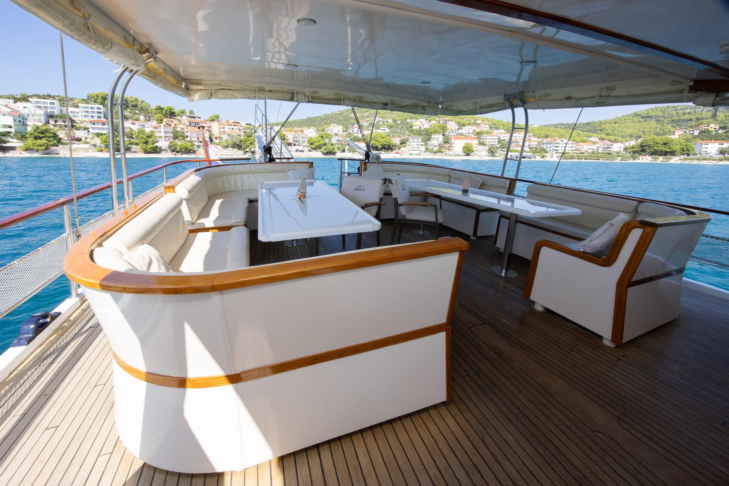 Gulet aft deck with white upholstered wraparound seating, teak-trimmed sofas, two dining tables, and canvas awning overhead