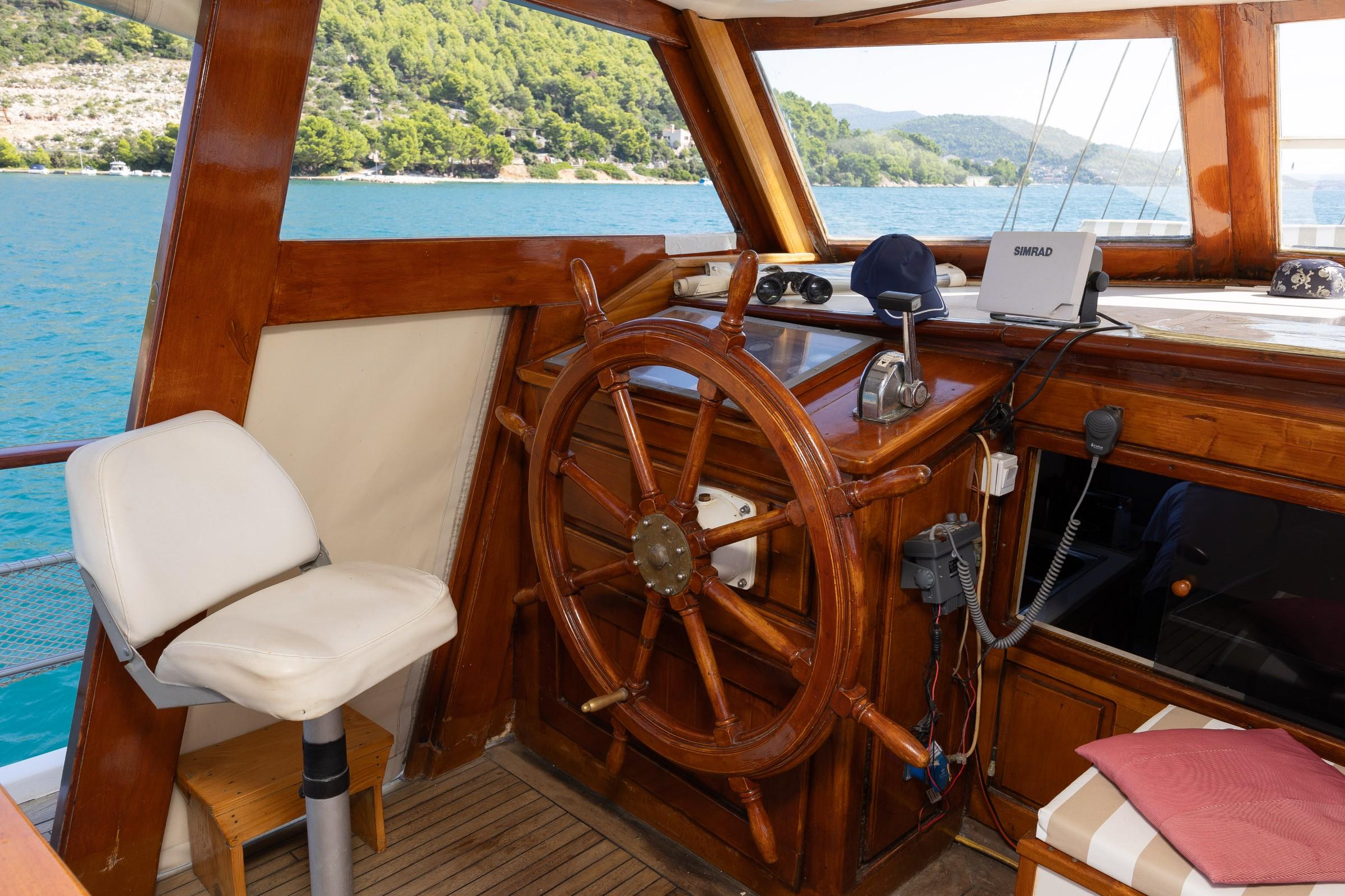 Traditional wooden gulet helm station with large brass-centred steering wheel, Simrad chartplotter, binoculars, and white captain's chair