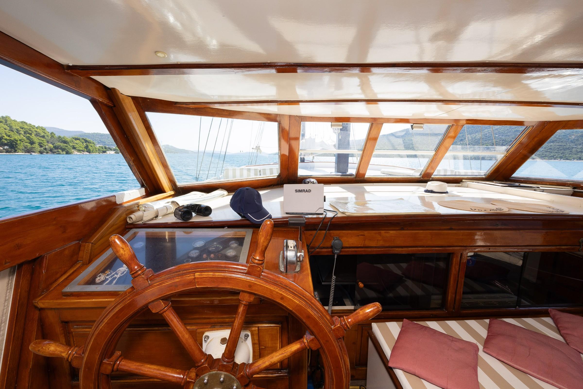 Classic gulet enclosed wheelhouse with varnished wooden helm wheel, Simrad navigation display, binoculars, and panoramic windows overlooking turquoise bay