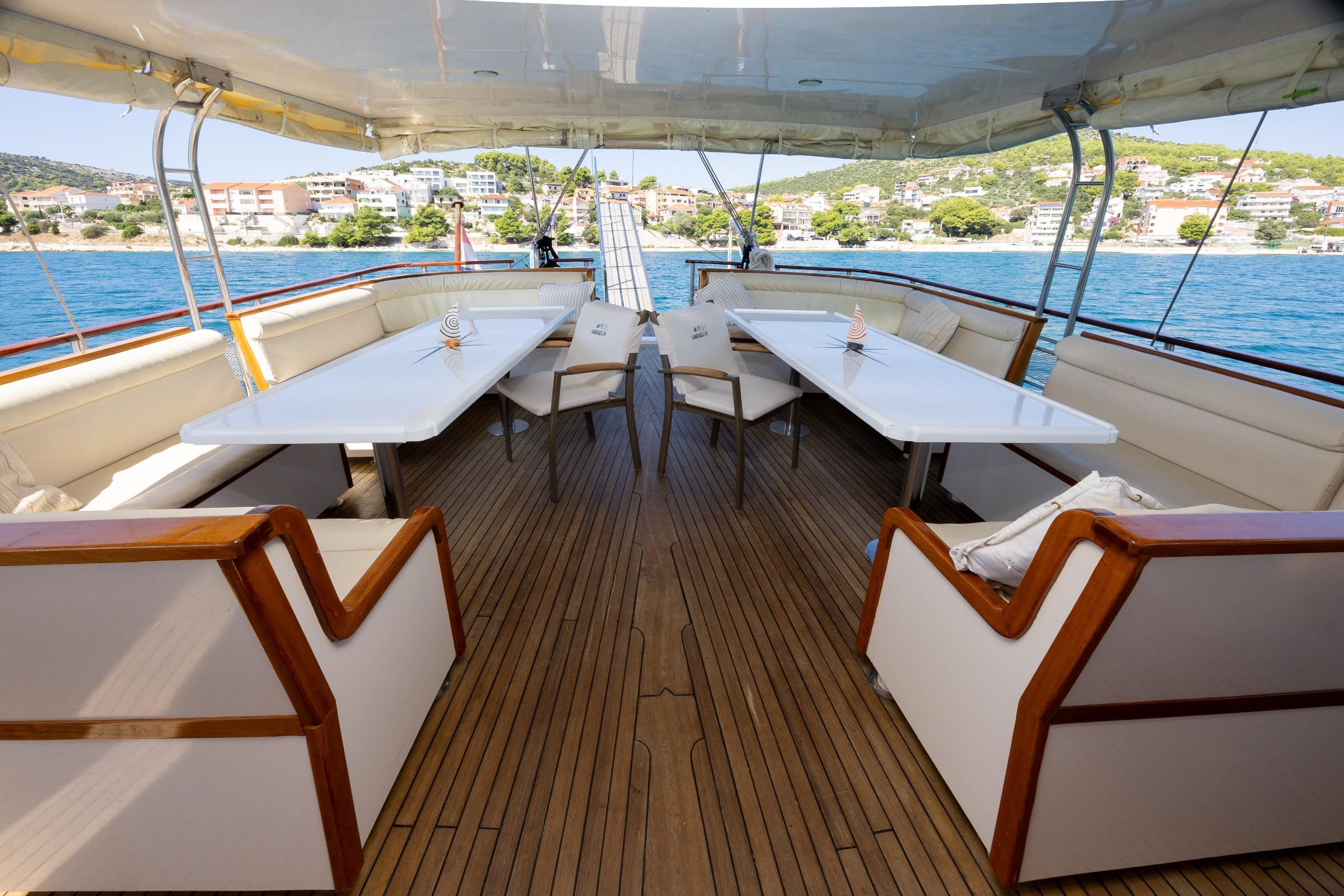 Gulet aft deck with two white dining tables, cream cushioned bench seating, teak wood chairs, and canopy overhead overlooking calm blue sea and coastal town