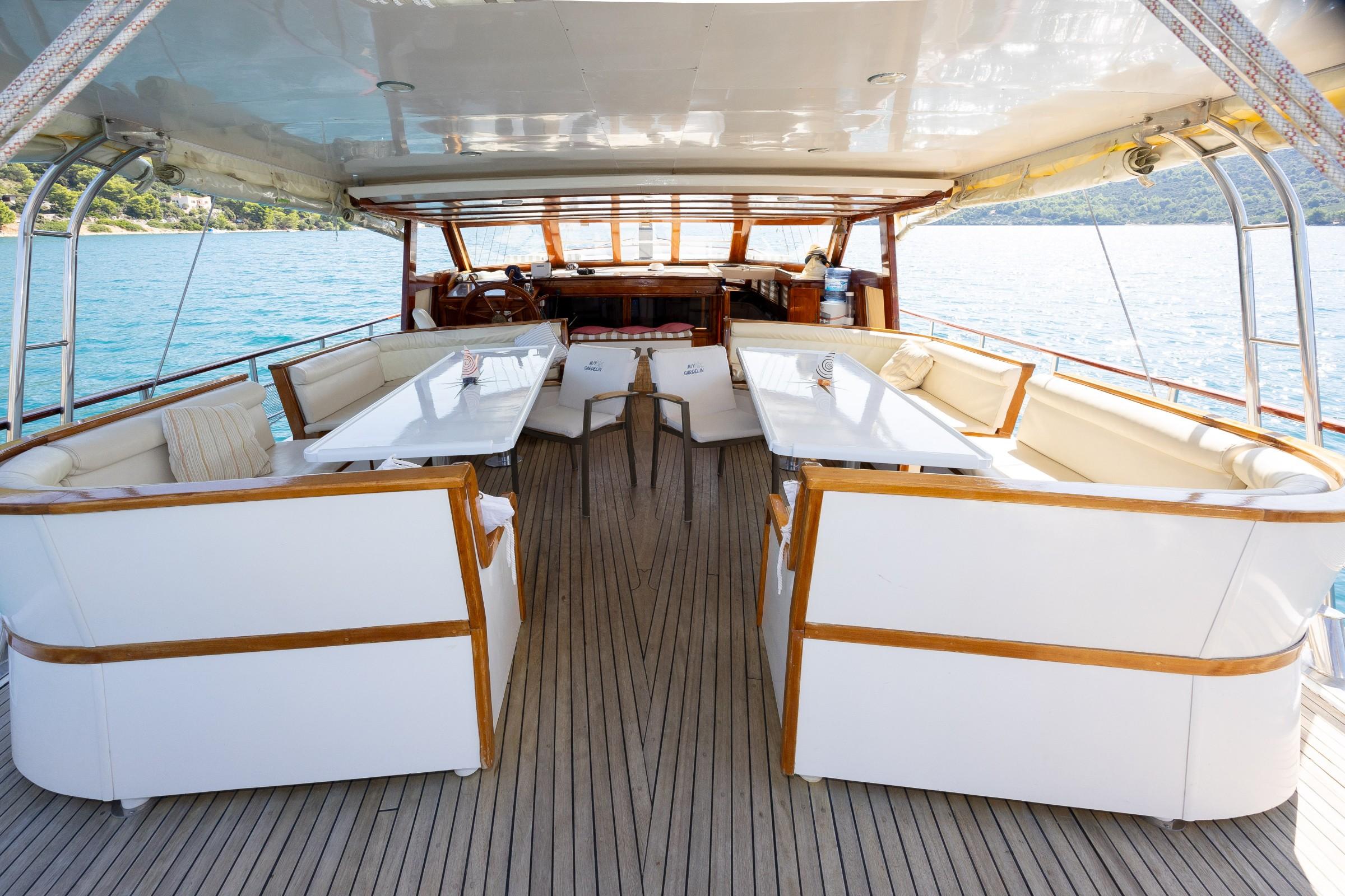 Gulet aft deck with twin white dining tables, wraparound cream sofas, teak decking and cockpit helm beyond