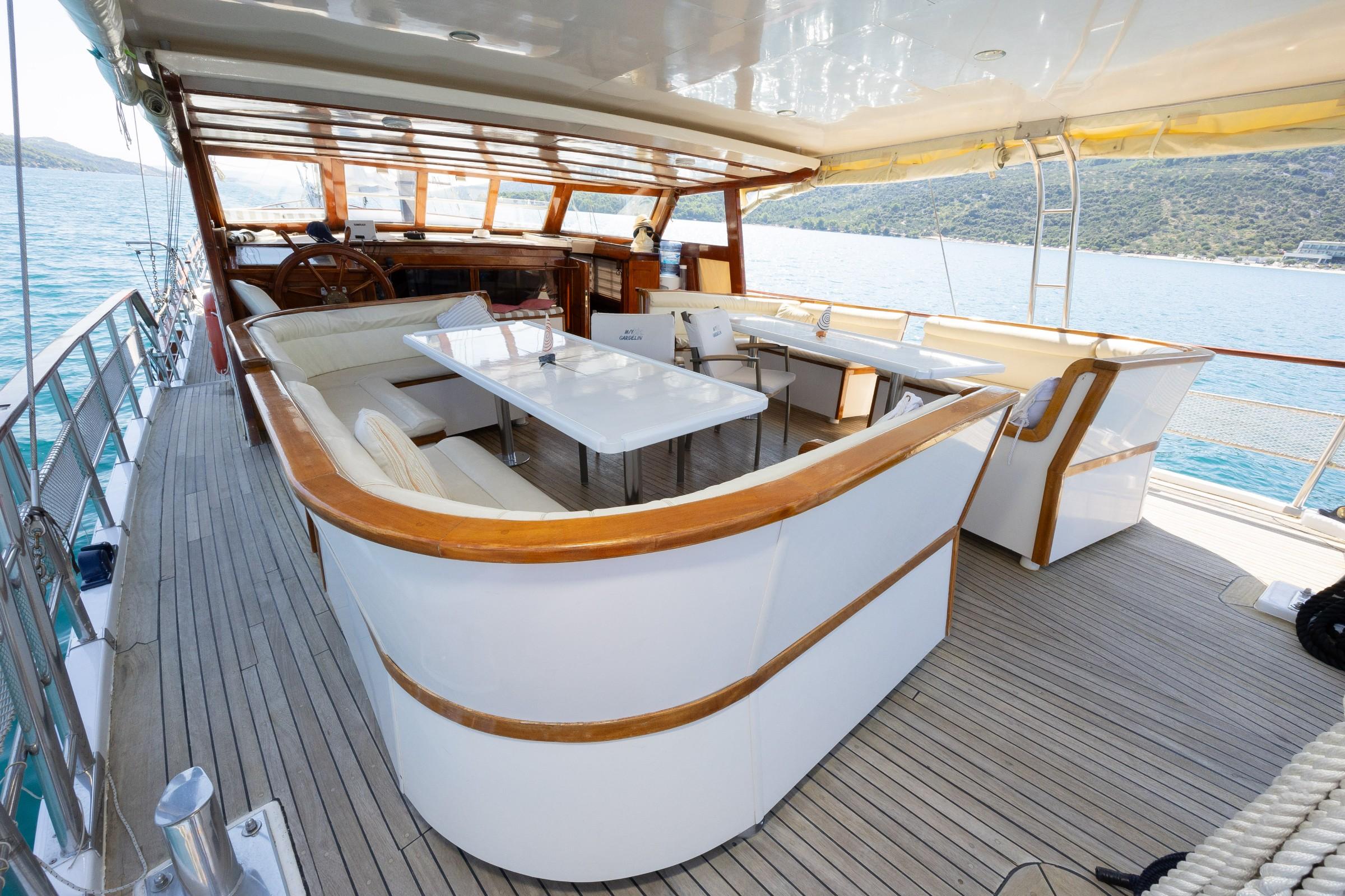 Gulet aft deck featuring curved white U-shaped seating, rectangular dining table, teak decking, and helm station behind