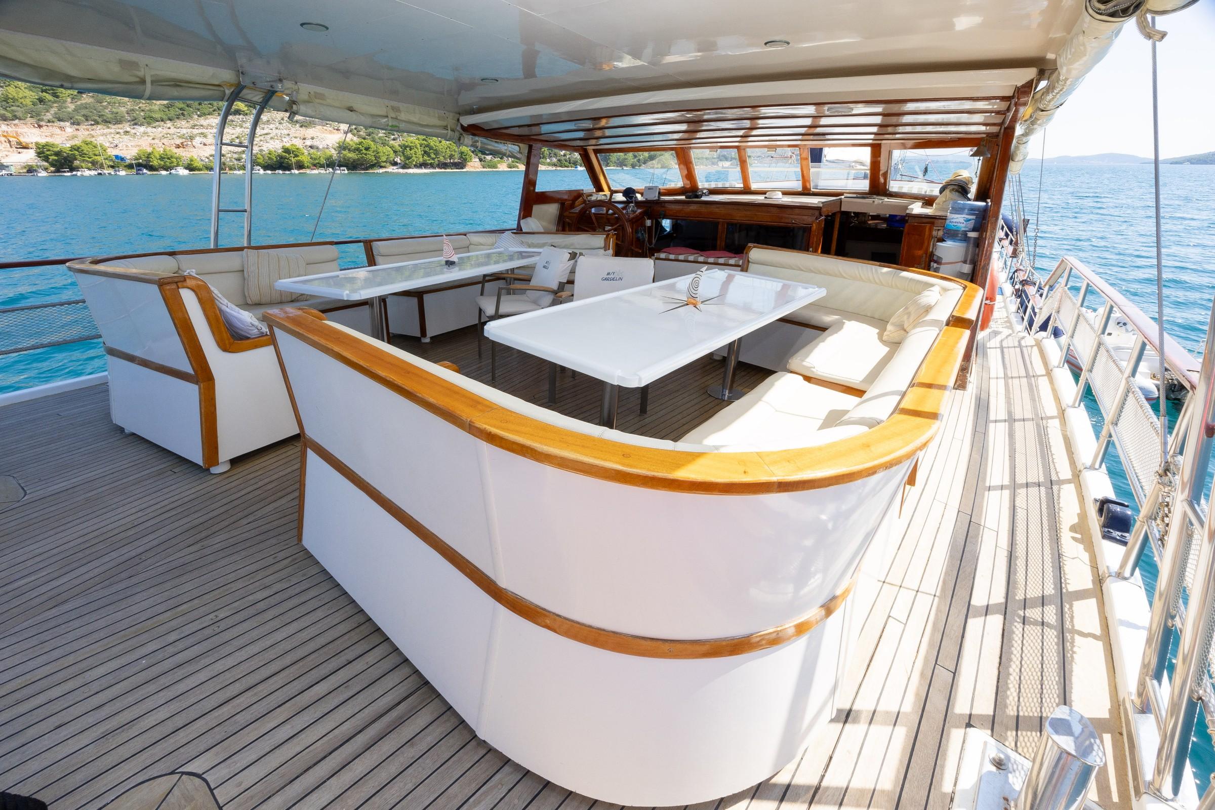 gulet aft deck with white wraparound bench seating, two white dining tables, teak decking, and helm station beyond
