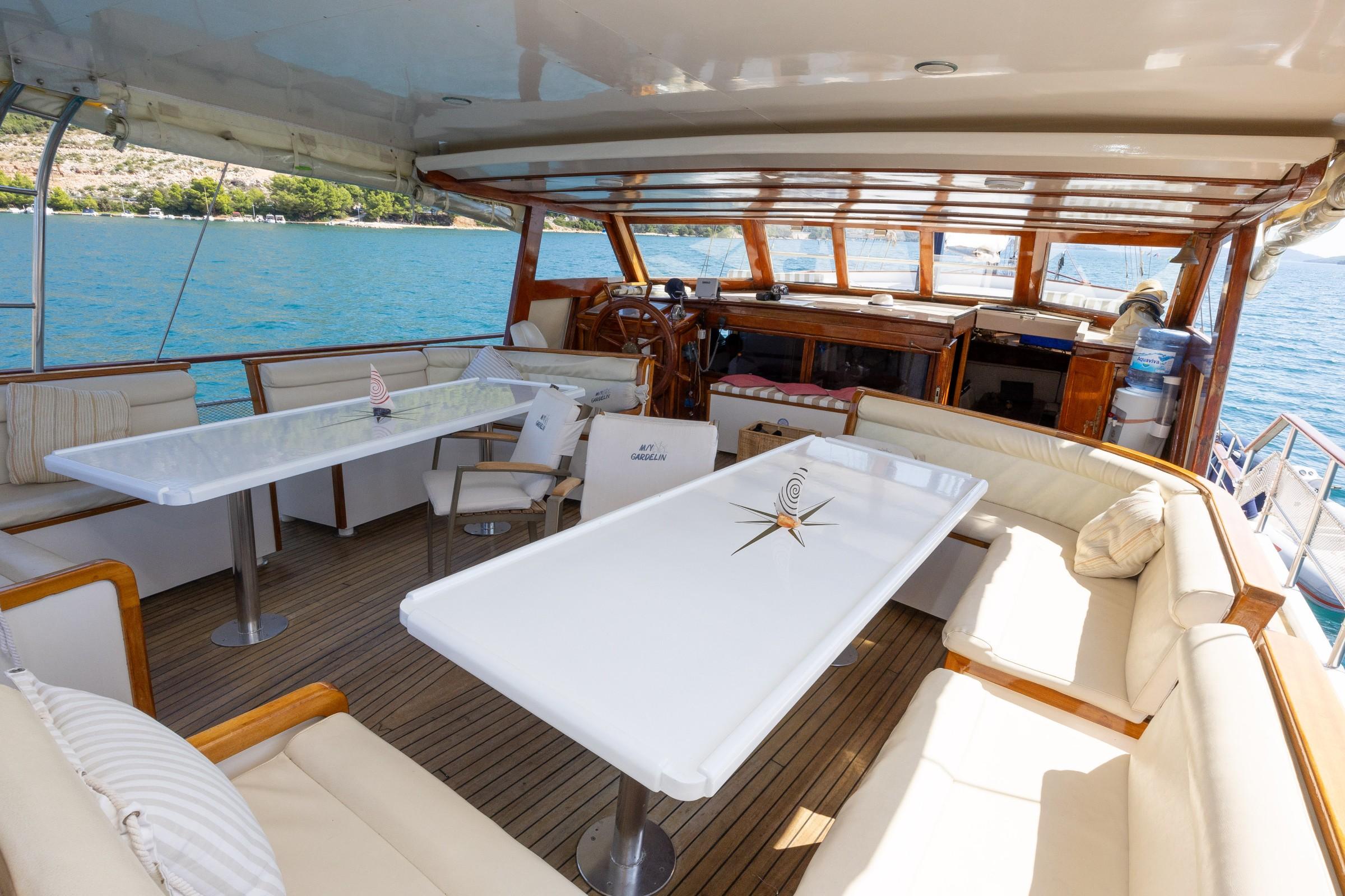 MV Gardelin gulet aft deck with cream cushioned seating, two white pedestal dining tables, teak decking, and helm station