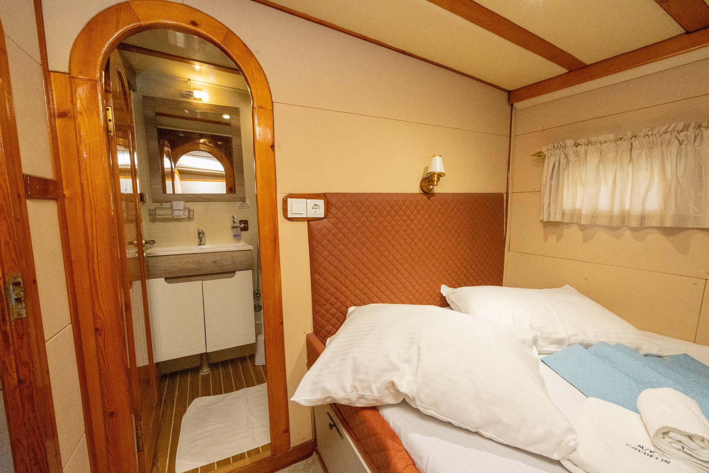 Gulet twin cabin with quilted terracotta headboard, white bedding, brass wall sconce, and arched wood-framed en-suite bathroom with vanity unit