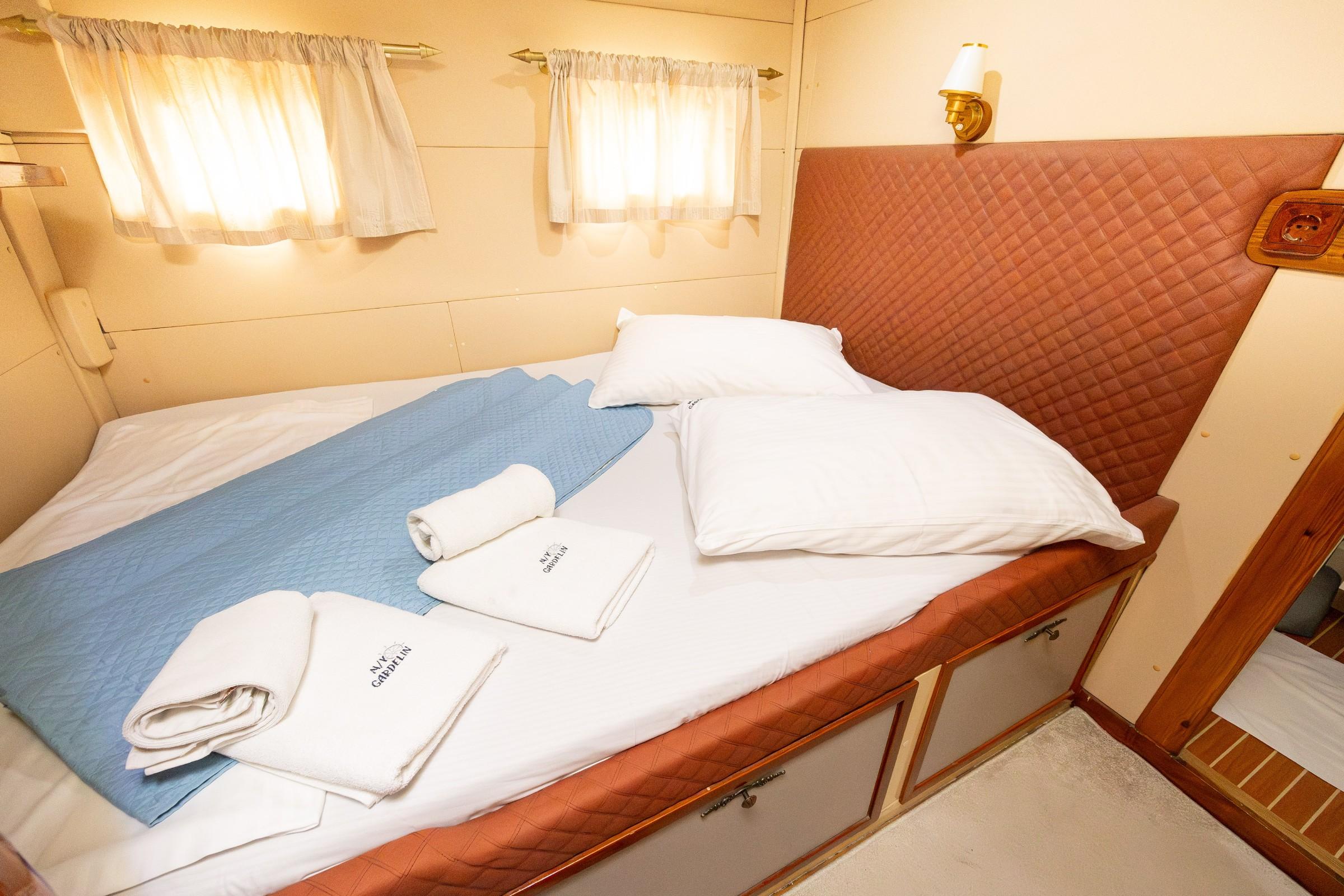 Gulet double cabin with terracotta quilted headboard, blue bedspread, white pillows, and branded towels laid out
