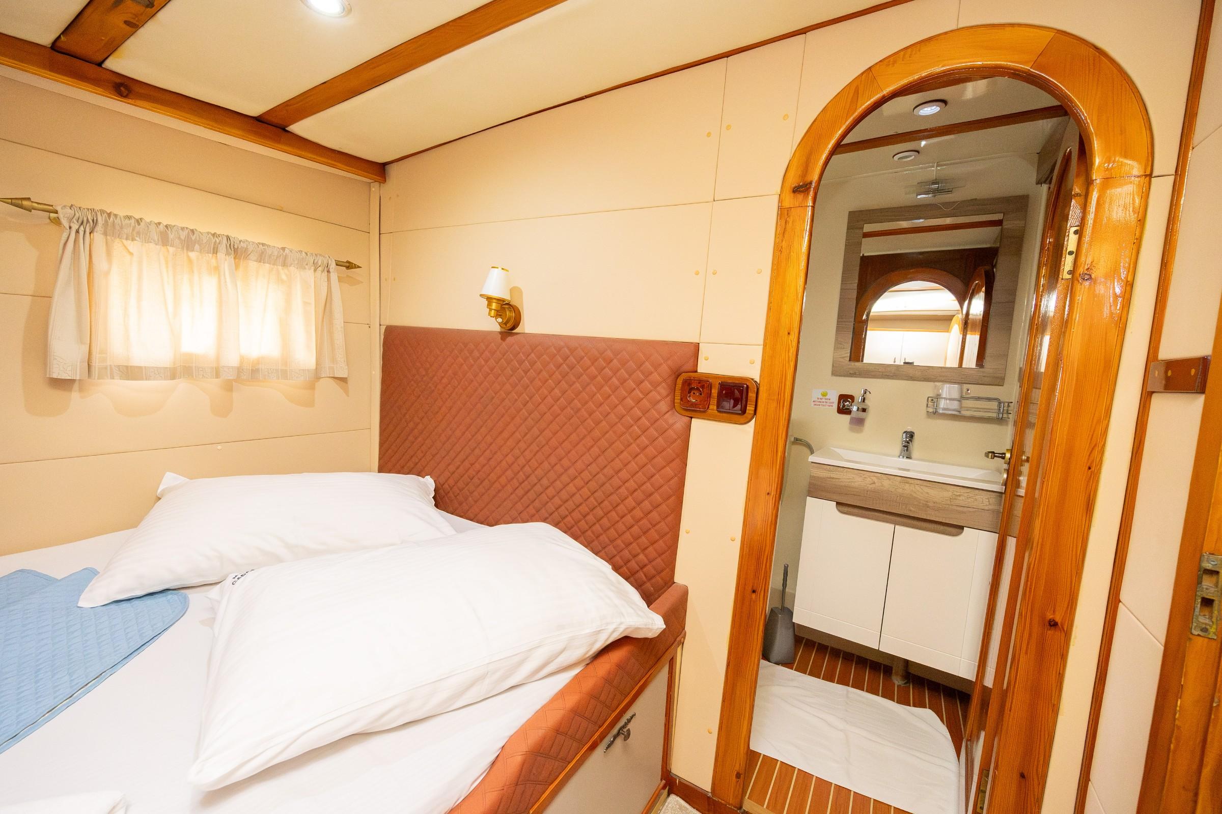 Gulet guest cabin with quilted terracotta headboard, white bedding, brass wall sconce, and arched timber-framed en-suite bathroom with vanity unit