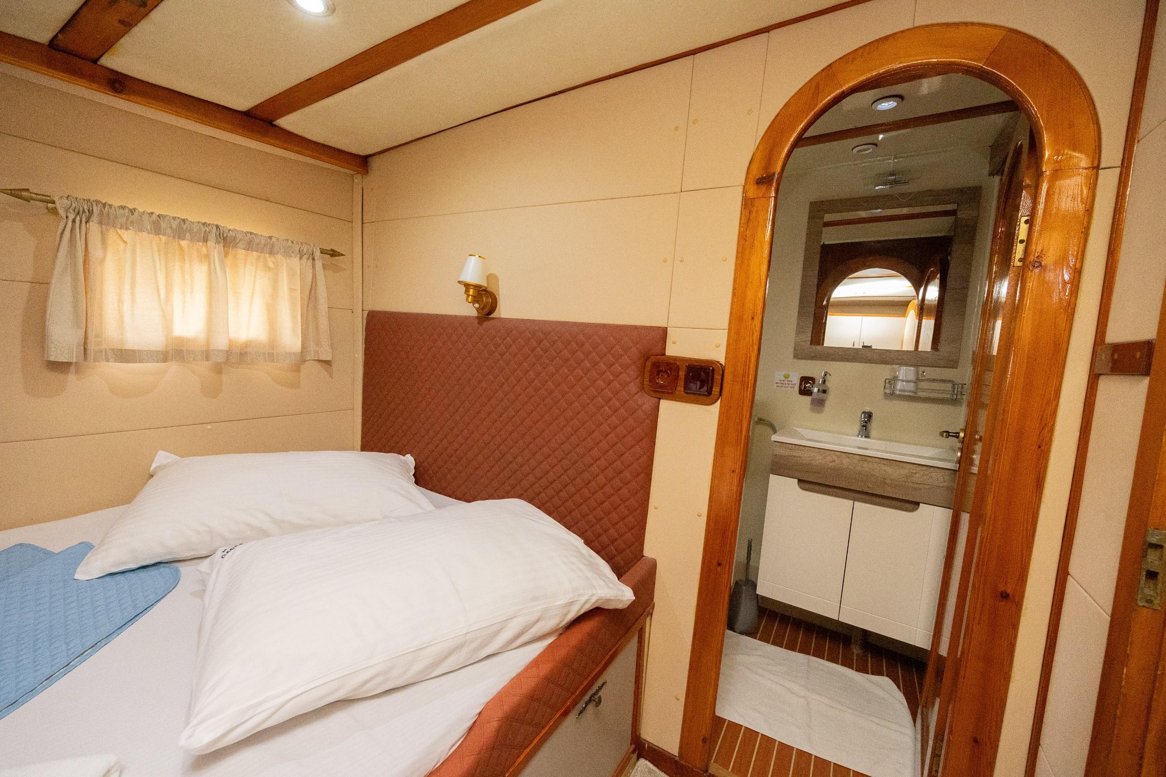 Gulet guest cabin with quilted red headboard, white bedding, wall sconce, and arched en-suite bathroom with vanity unit