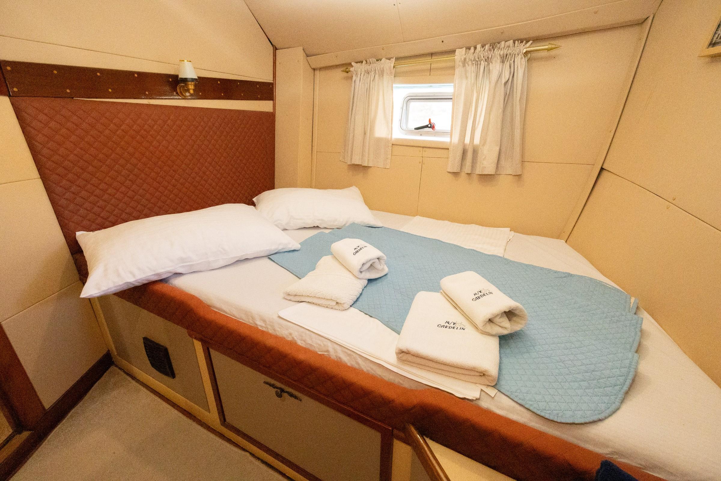 Double cabin aboard M/Y Gardelin with quilted blue runner, white pillows, branded towels, and curtained porthole window