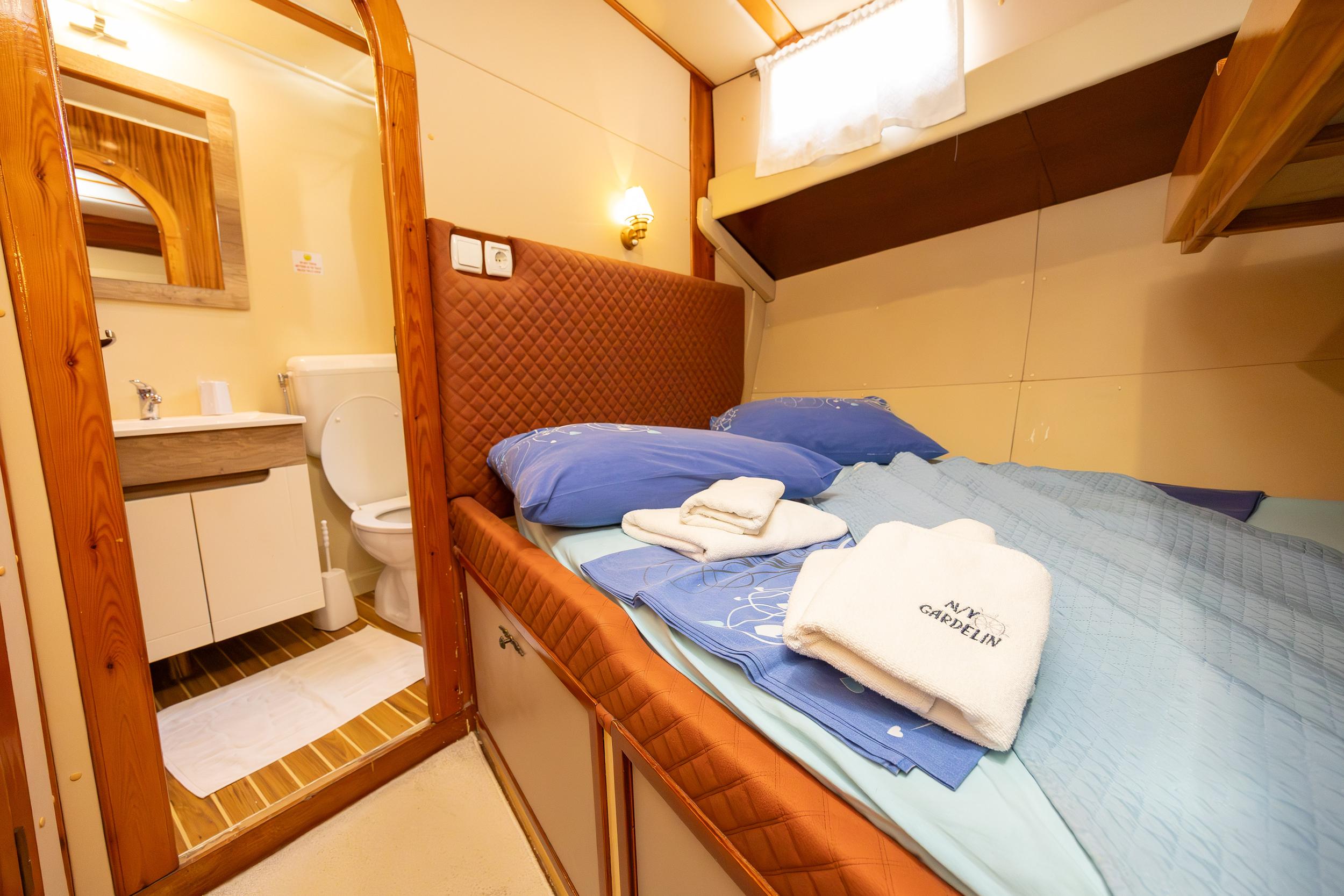 Gulet double cabin with quilted headboard, blue bedding, folded towels, and open en-suite showing toilet and sink