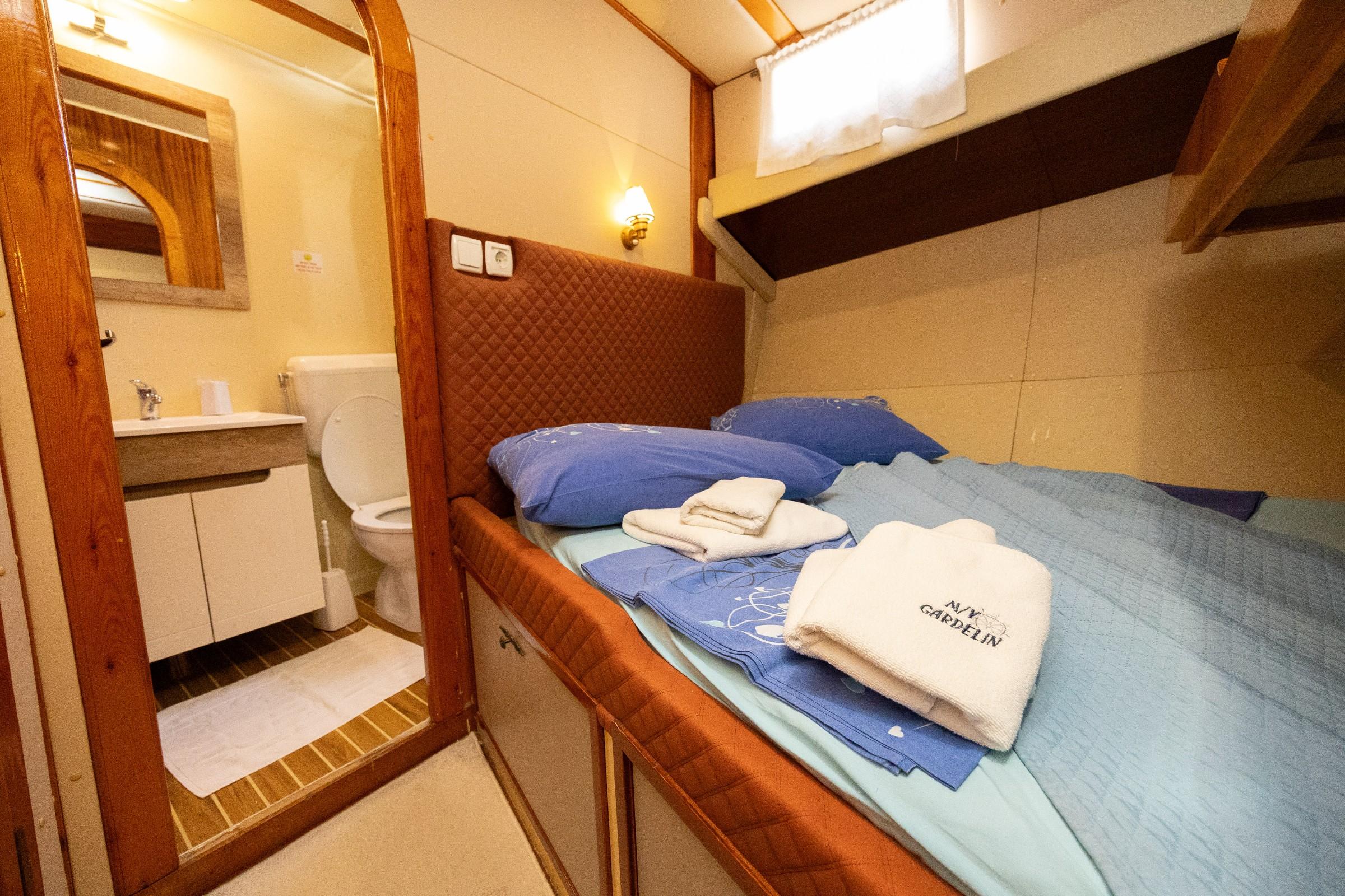 Gulet cabin with blue bedding, quilted headboard, folded MV Gardelin branded towels, and en-suite toilet and sink visible