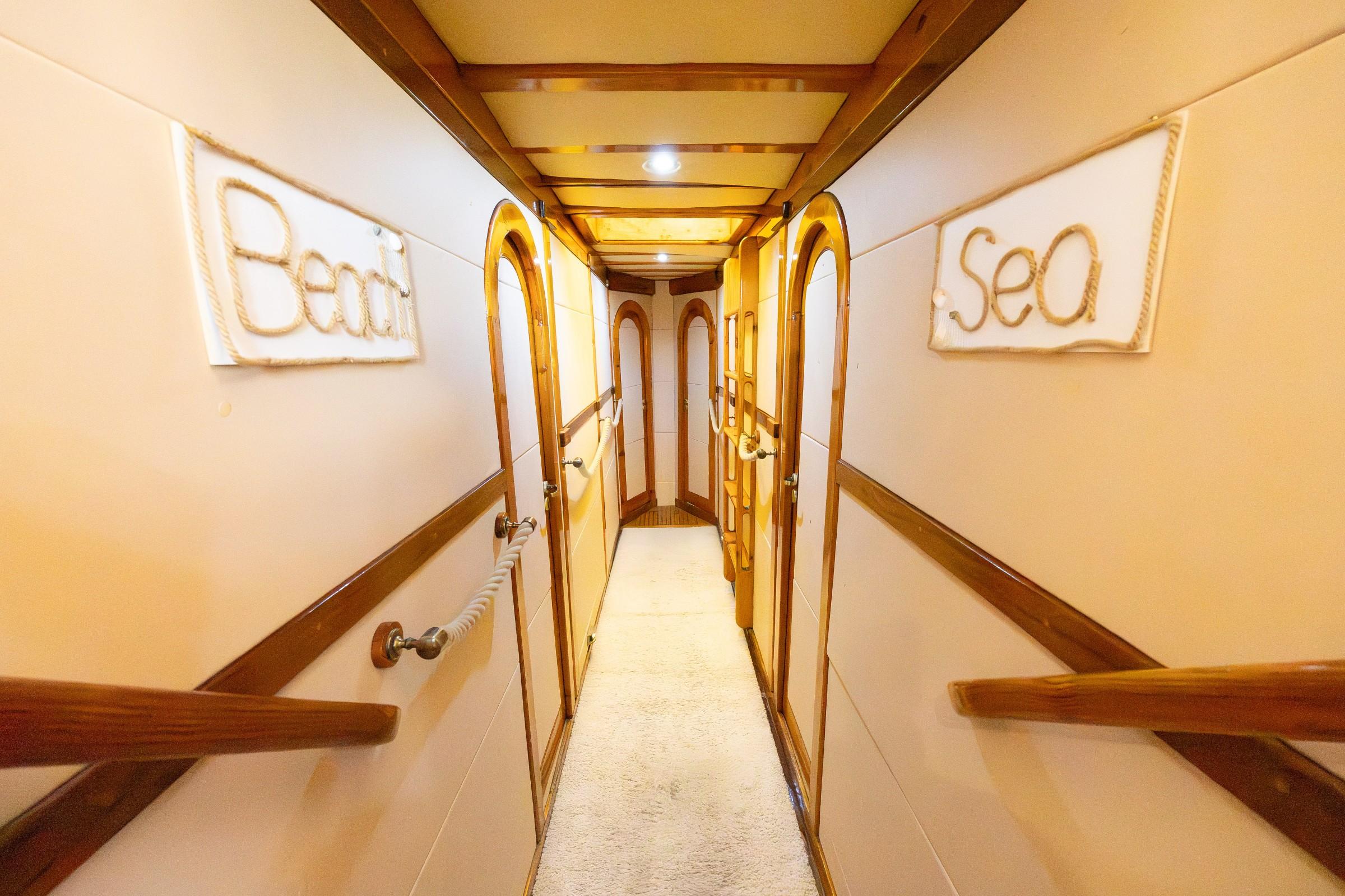 Below-deck corridor on luxury gulet with arched teak-framed cabin doors, rope handrails, cream carpet, and nautical rope wall signs reading Beach and Sea