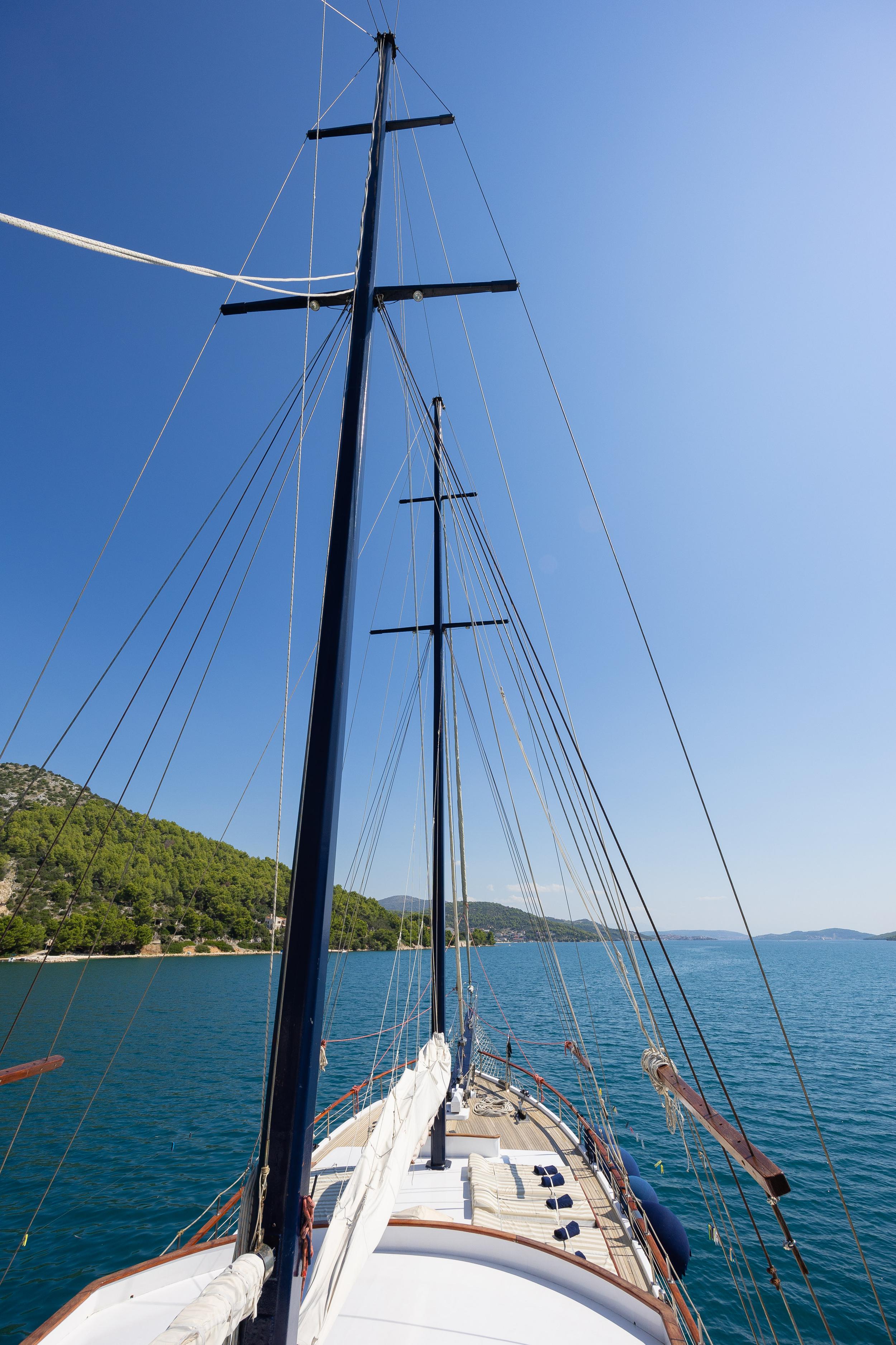 Gulet bow deck with twin dark masts, rigging, and cushioned sun loungers anchored in calm turquoise bay