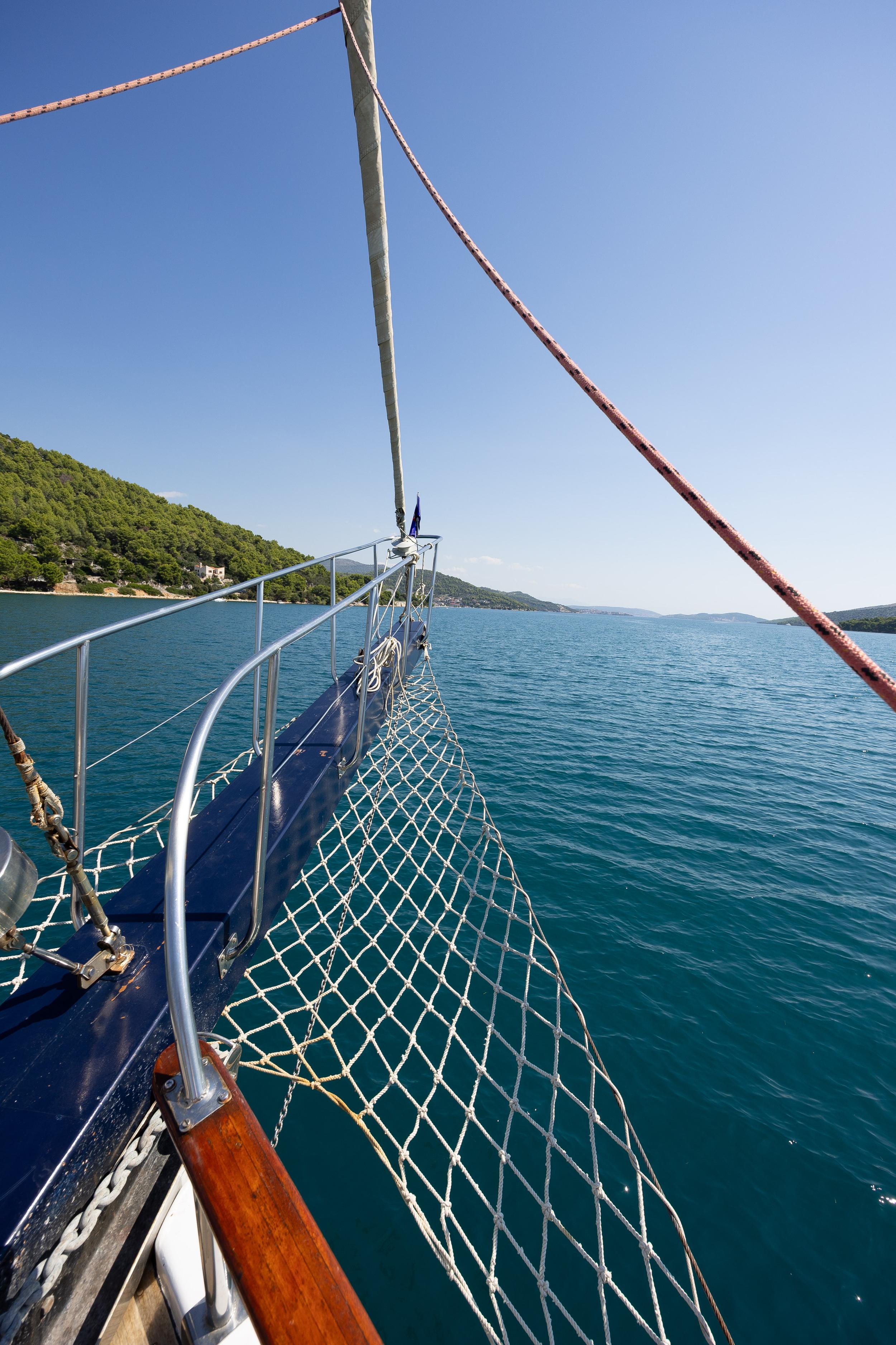 Gulet bow deck with rope safety net and stainless steel railings sailing calm turquoise waters past forested hillside