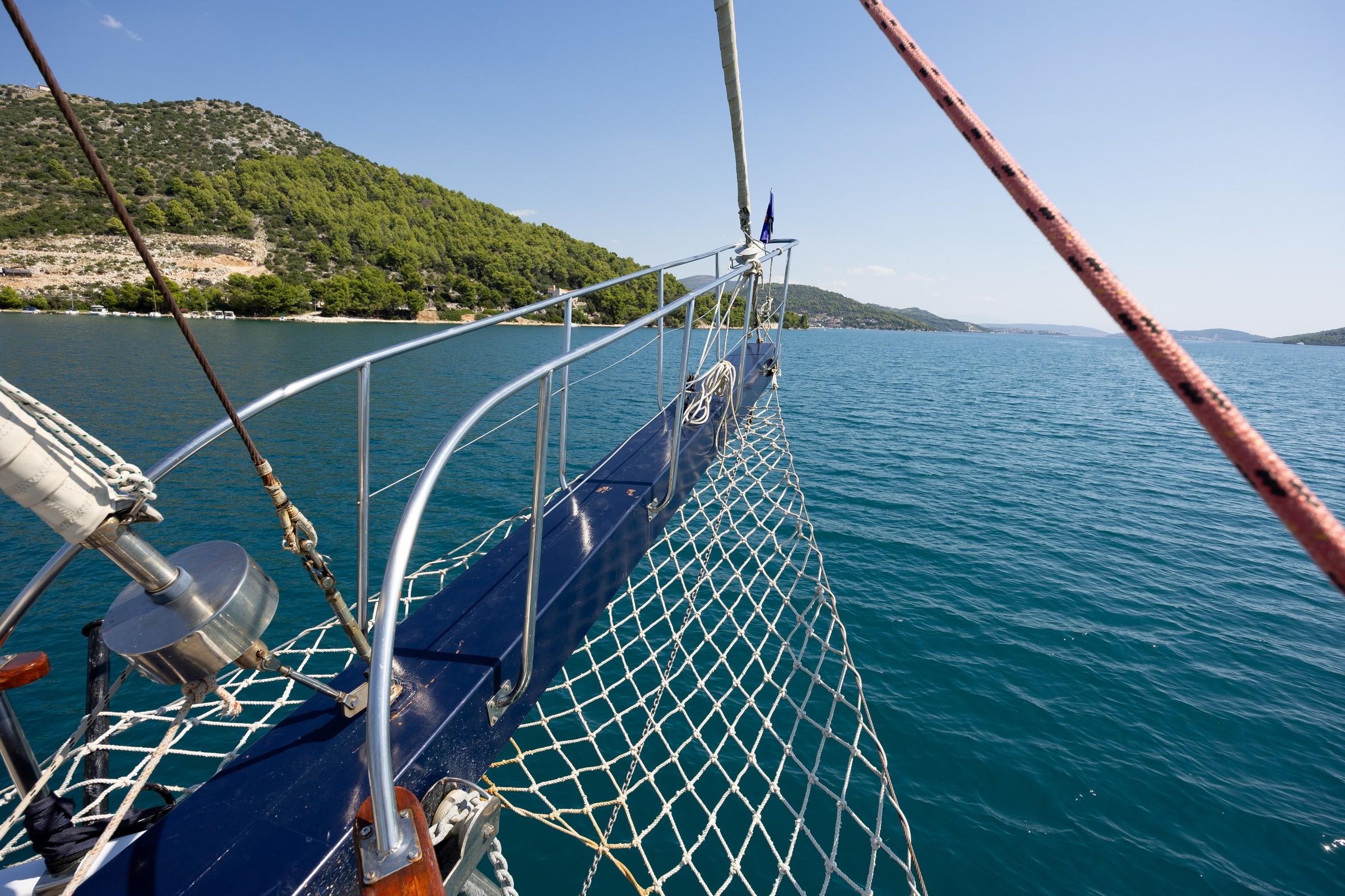 Gulet bow with white rope safety netting, polished steel bowsprit rail, and blue hull against calm turquoise Croatian waters