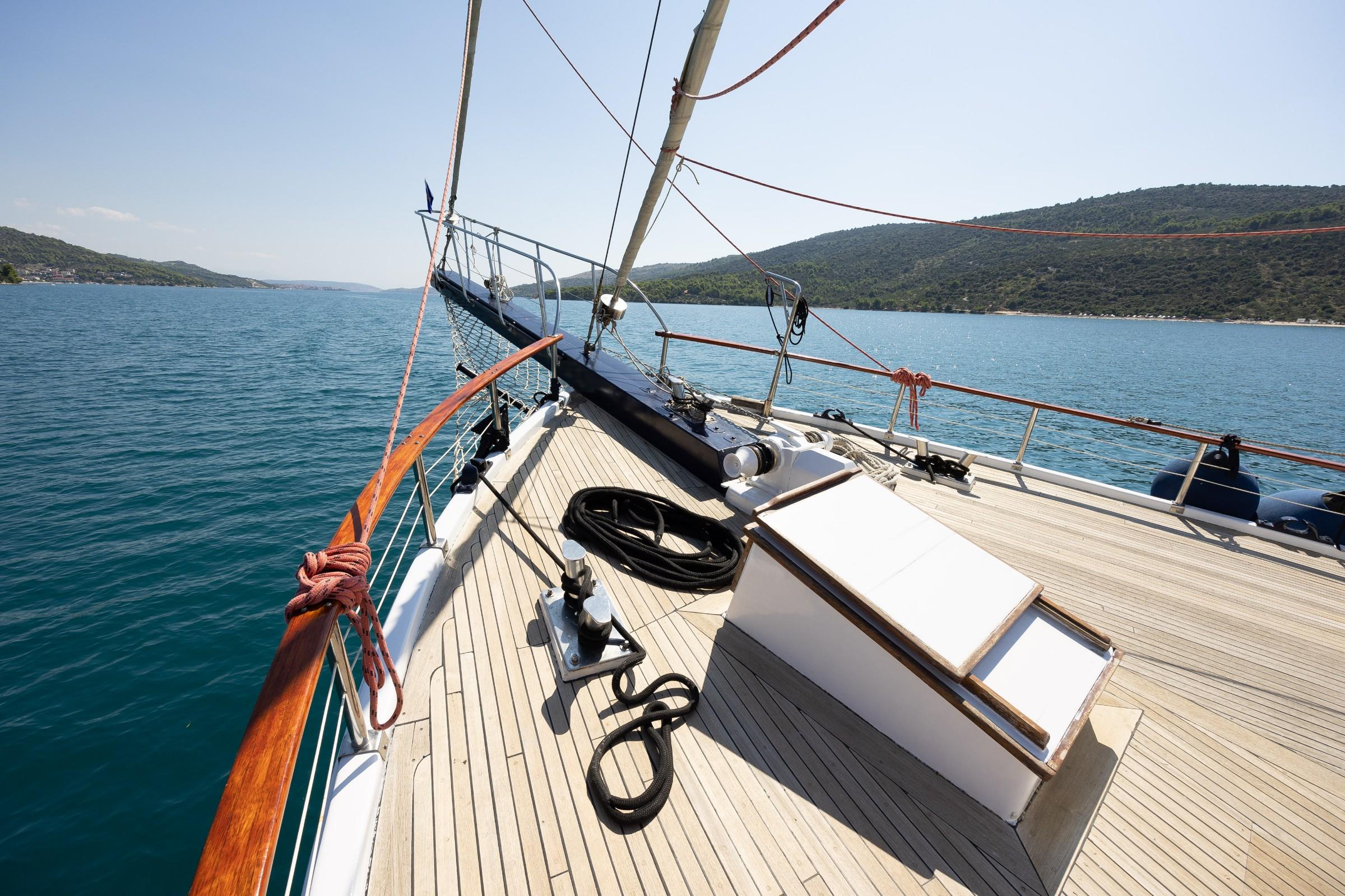 Gulet foredeck teak deck with mast rigging, mooring cleats, coiled lines and wooded hillside backdrop