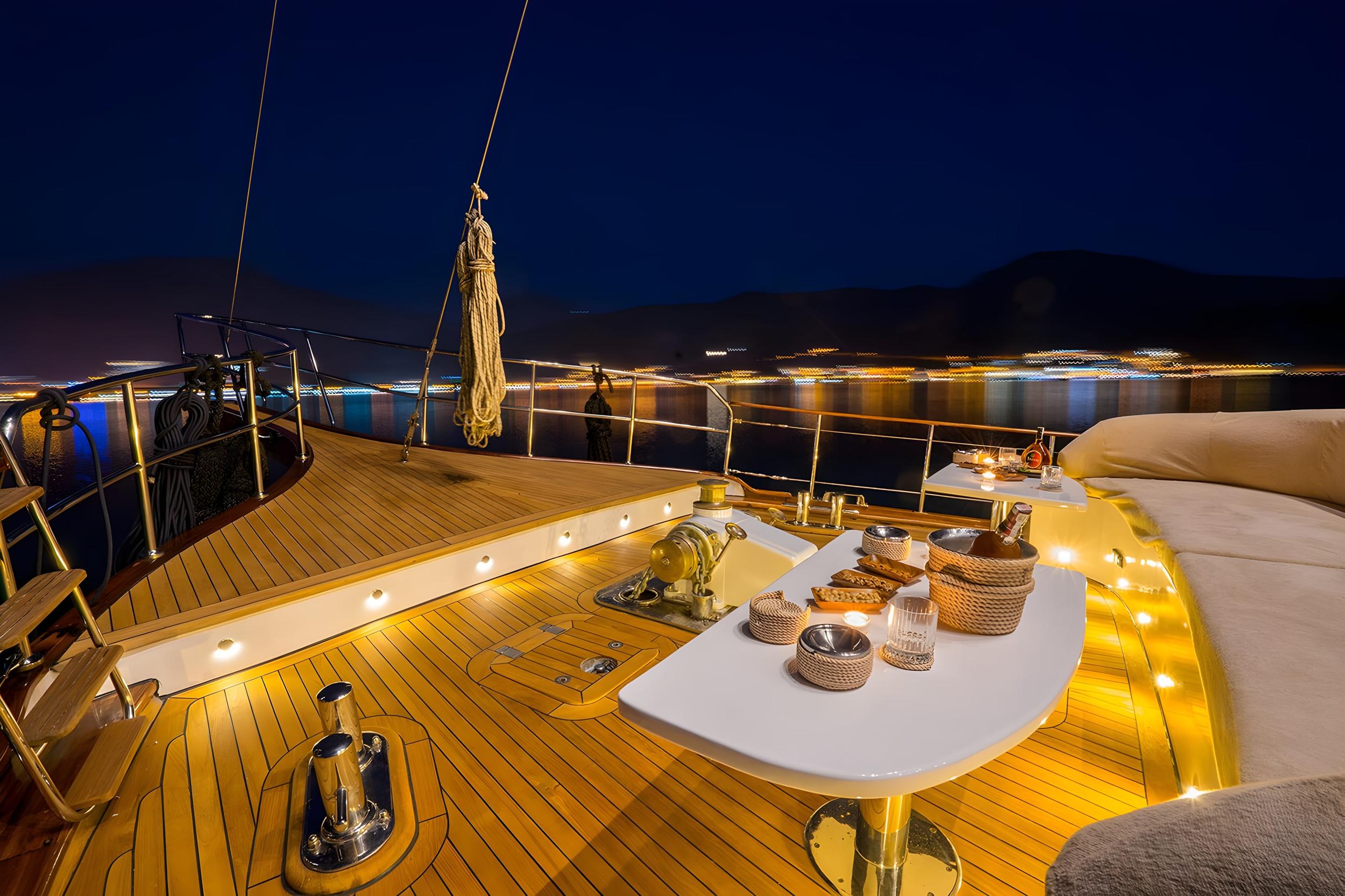 gulet foredeck teak deck at night with white pedestal table set with snacks, champagne bucket, candle and glassware under warm deck lighting