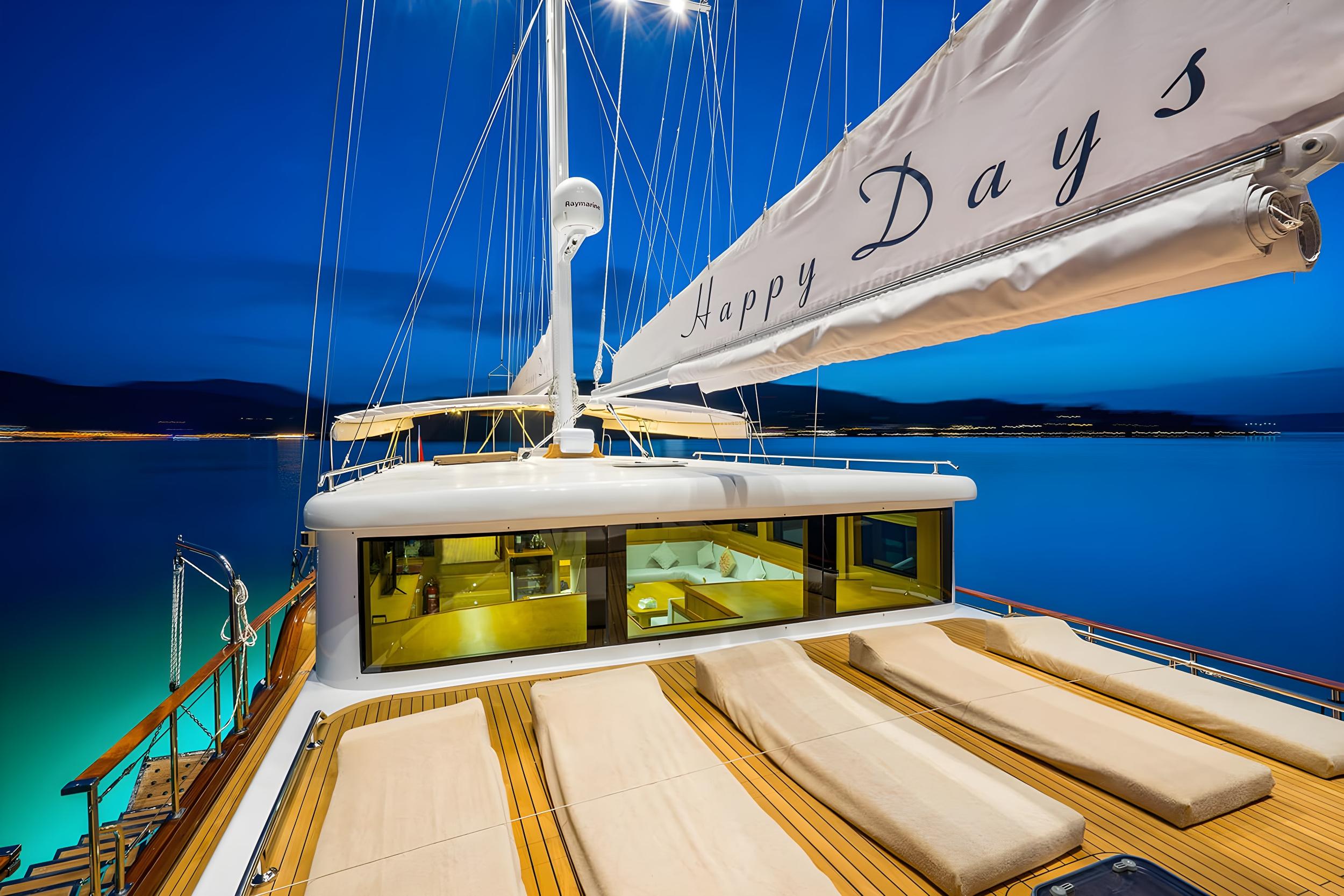 Gulet Happy Days foredeck teak deck with cream sun loungers and glowing salon interior at night