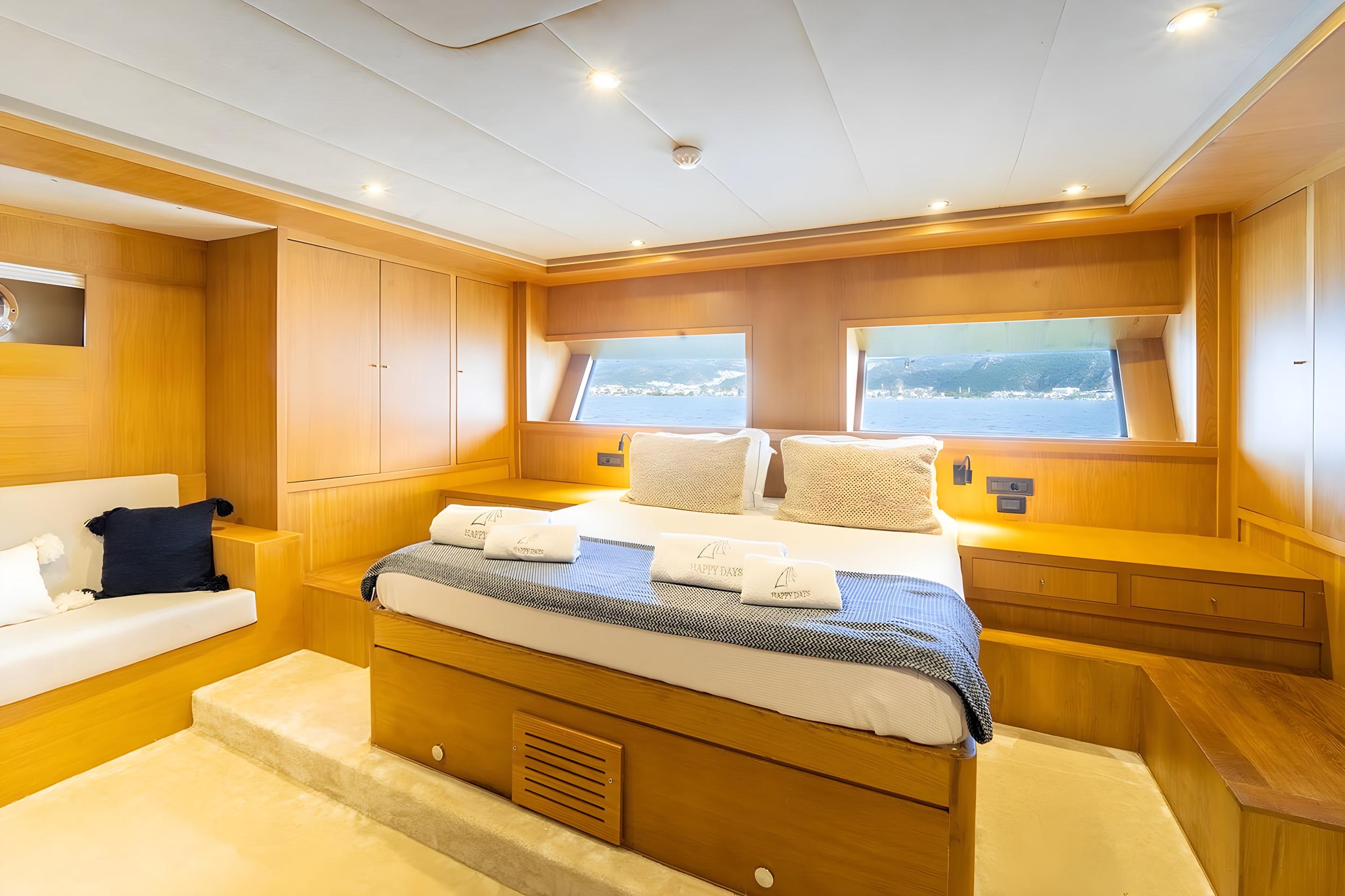 Master cabin on gulet Happy Days featuring king-size bed with branded towels, built-in wood cabinetry, and dual panoramic windows overlooking the sea
