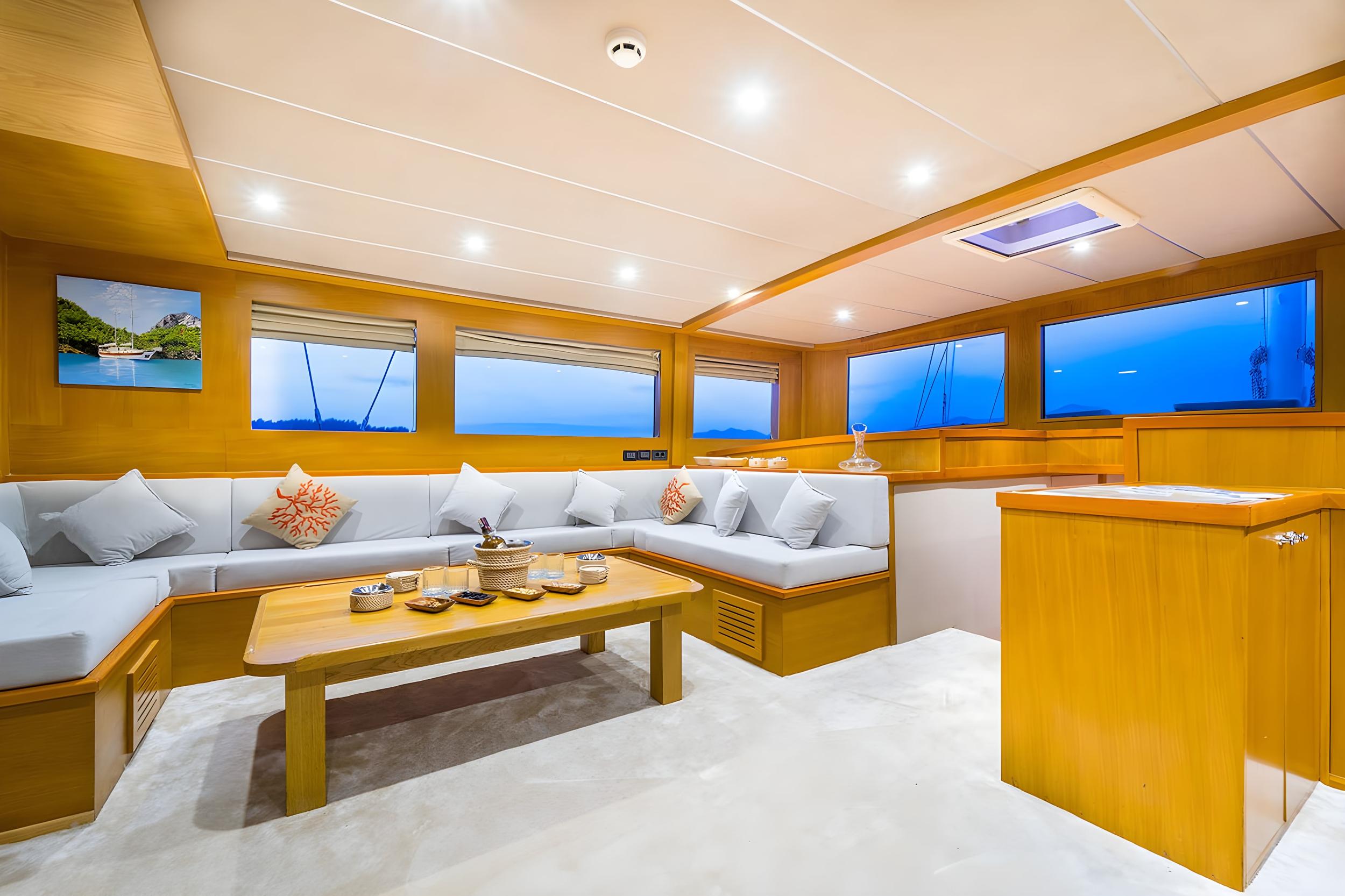 Gulet main saloon with white L-shaped seating, teak coffee table, and panoramic windows at dusk
