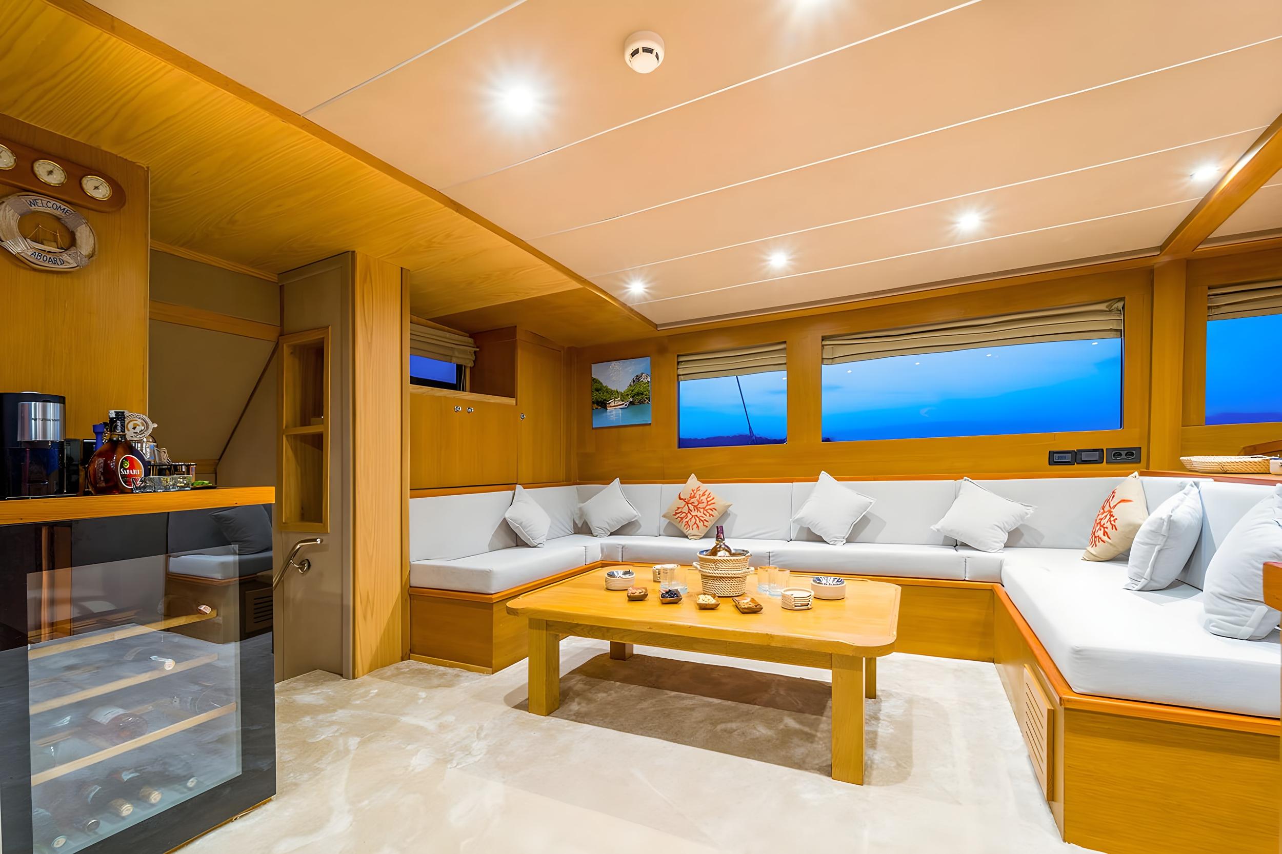 Gulet indoor salon with L-shaped white cushioned sofas, teak coffee table, and glass wine cooler stocked with bottles