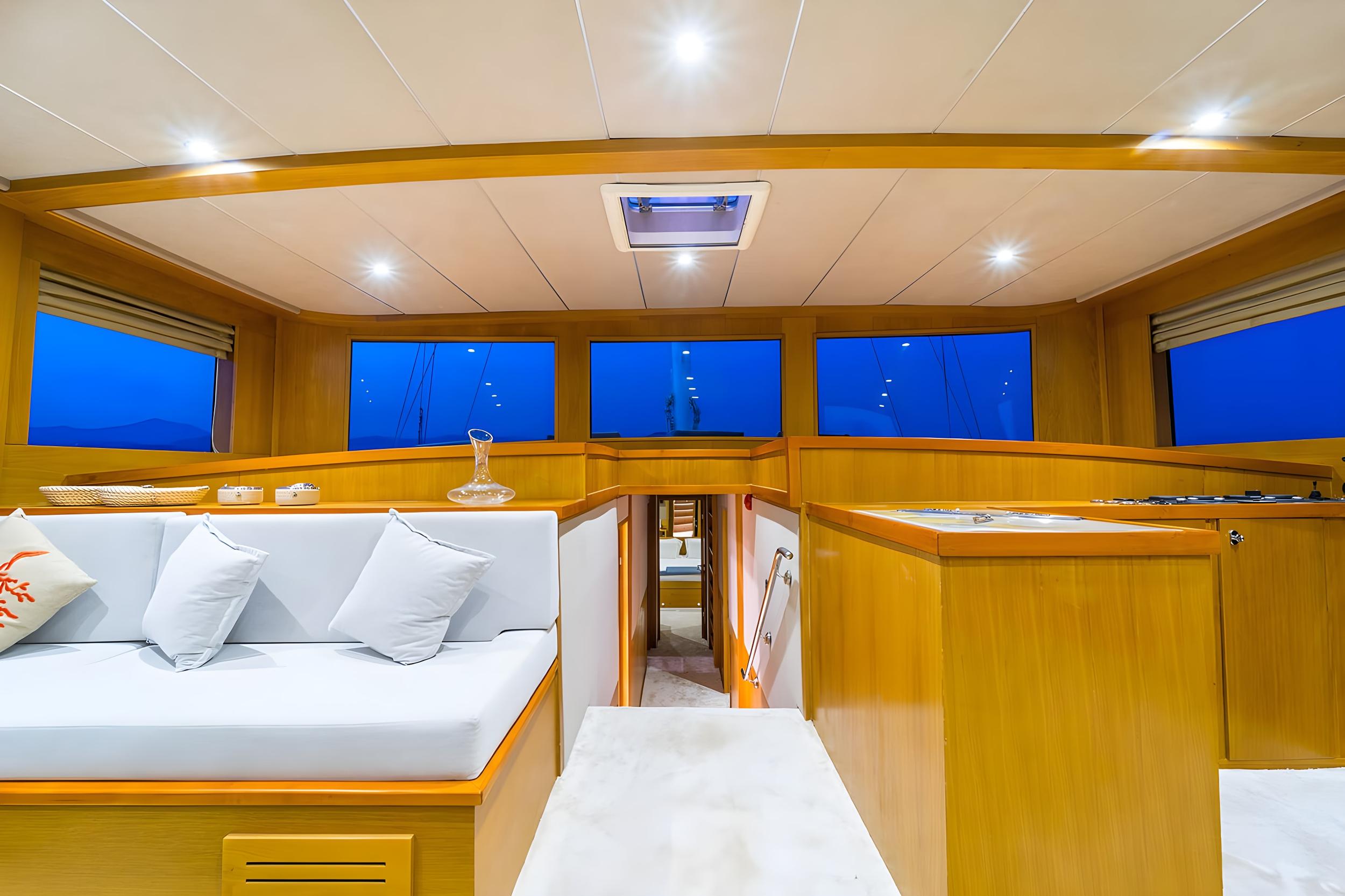 Gulet interior saloon with white upholstered seating, honey-toned wood panelling, galley counter, and panoramic windows at dusk