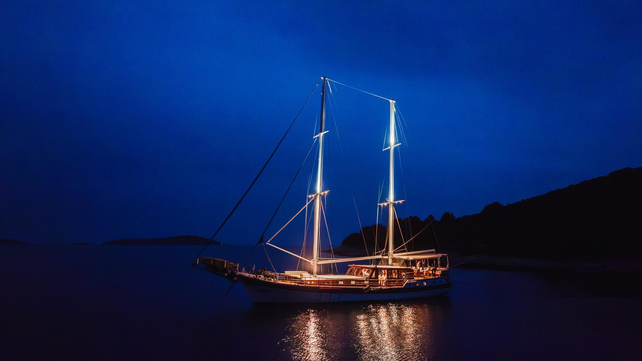 Traditional wooden gulet yacht illuminated at night anchored in calm bay with dark hills