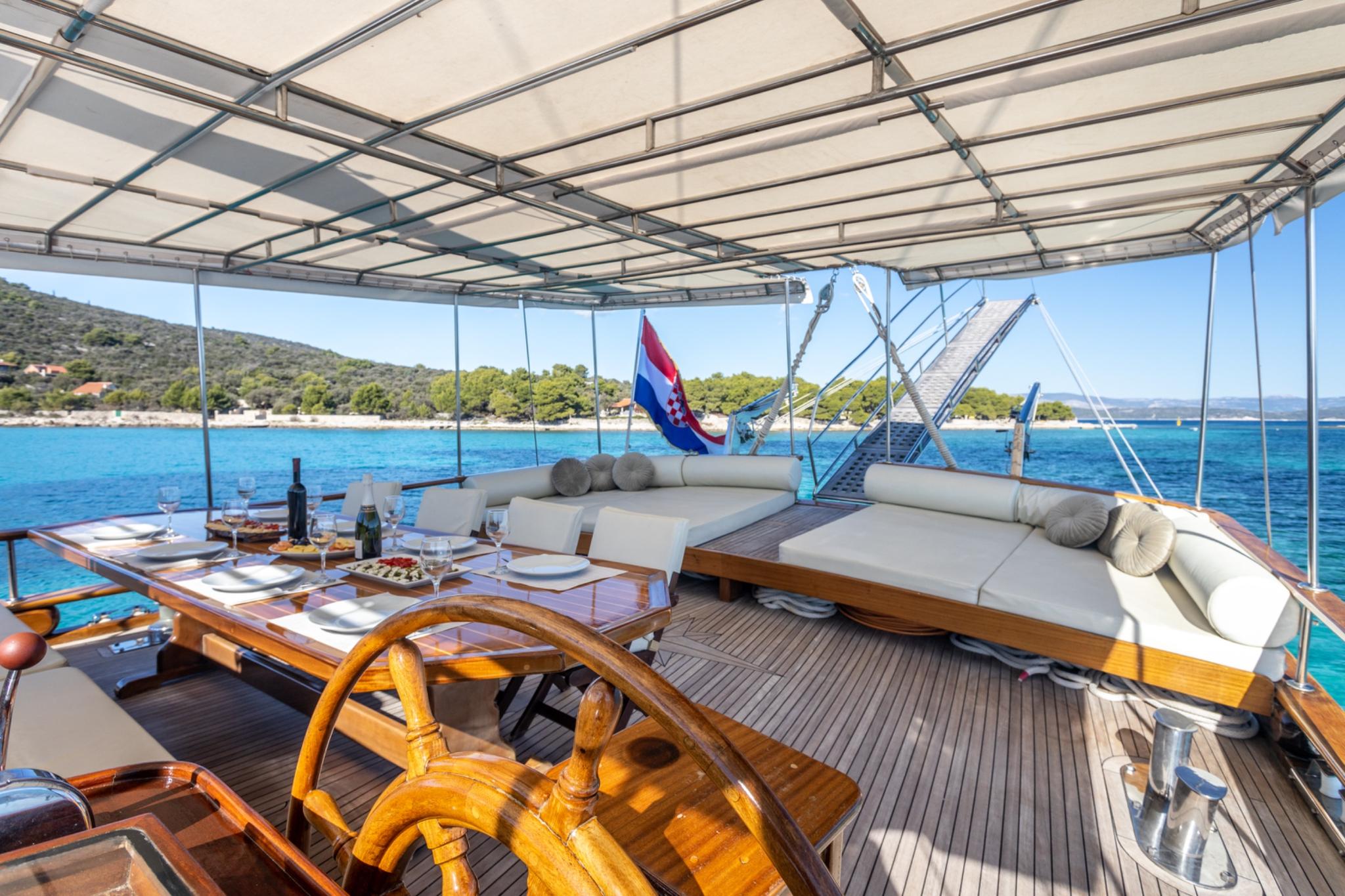 gulet aft deck with wooden dining table, white cushioned seating and Croatian flag