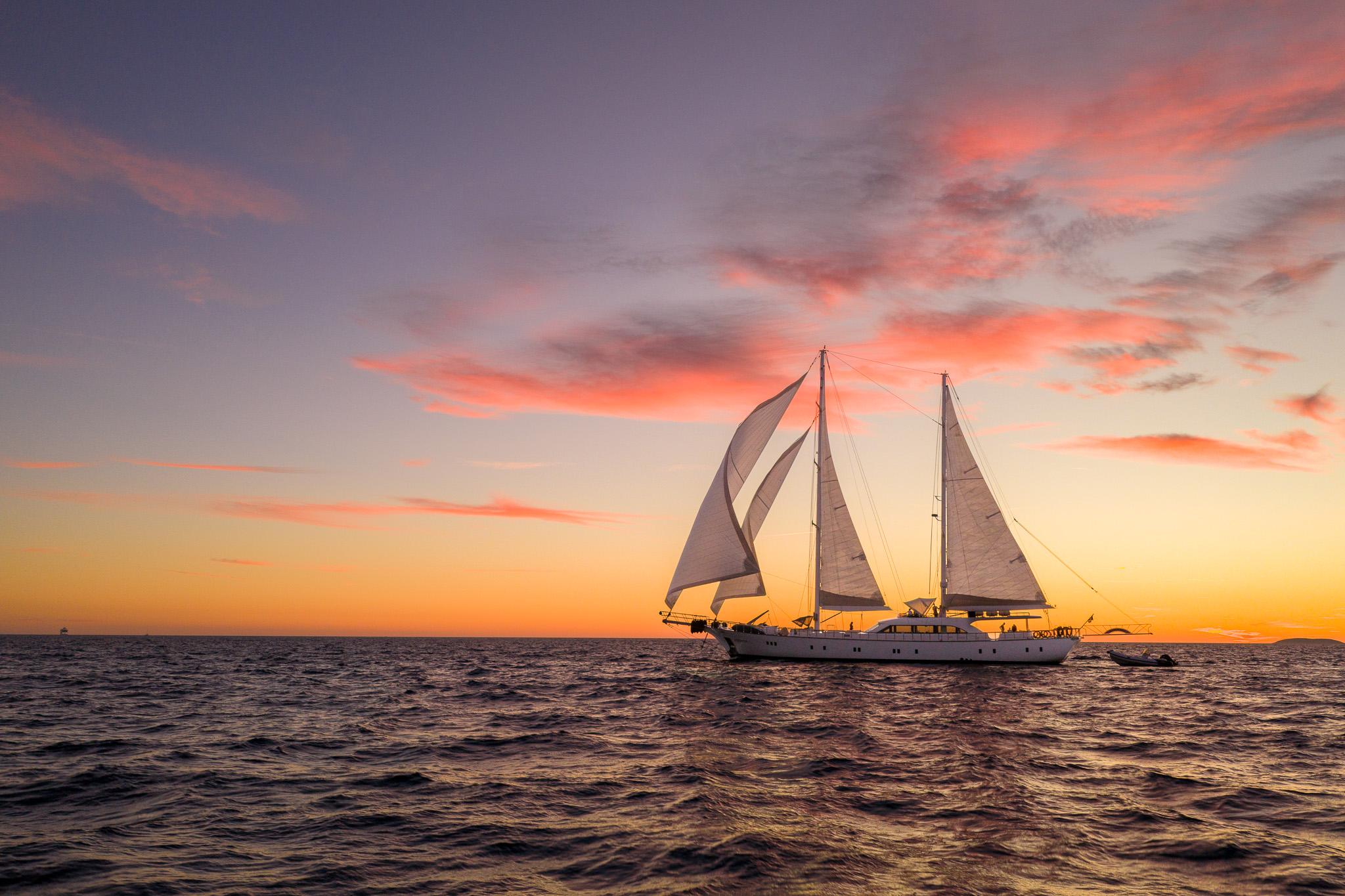 White sailing gulet with multiple sails deployed at sunset on calm waters