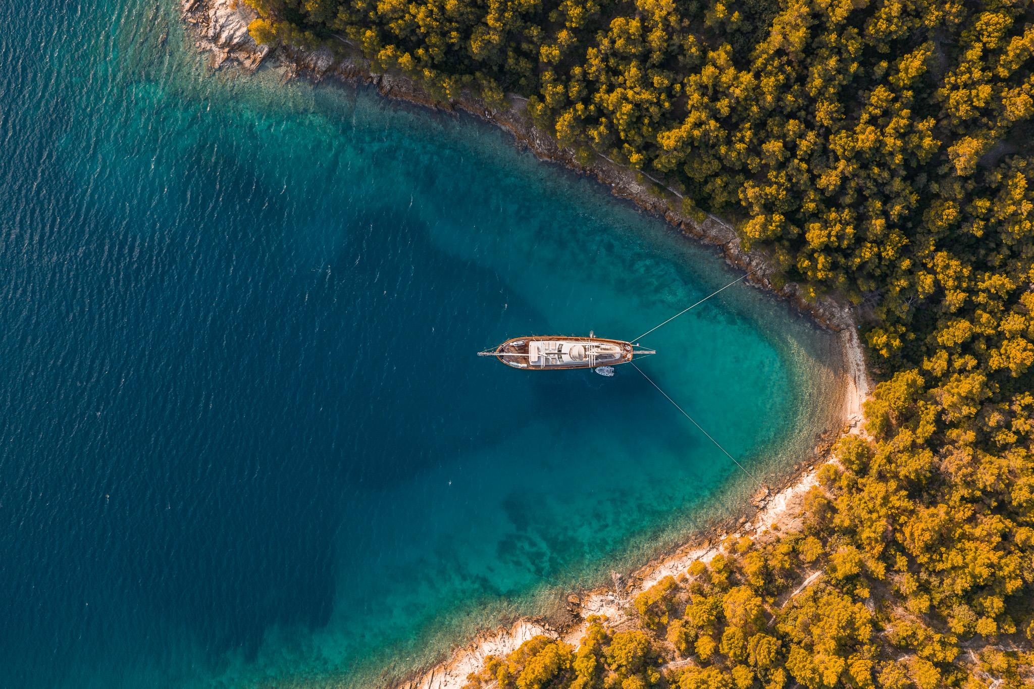 aerial view of traditional wooden gulet yacht anchored in crystal clear turquoise bay surrounded by golden autumn forest