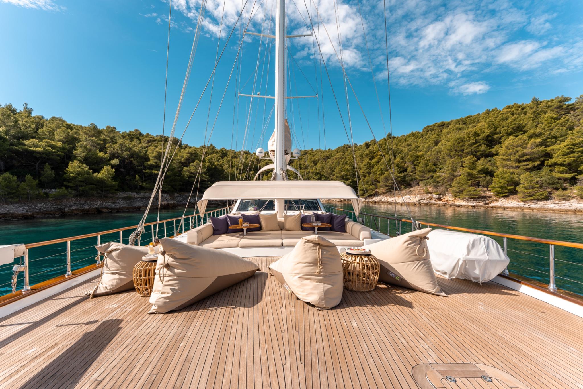 Gulet foredeck with beige cushioned seating, teak decking, and rigging in sheltered bay