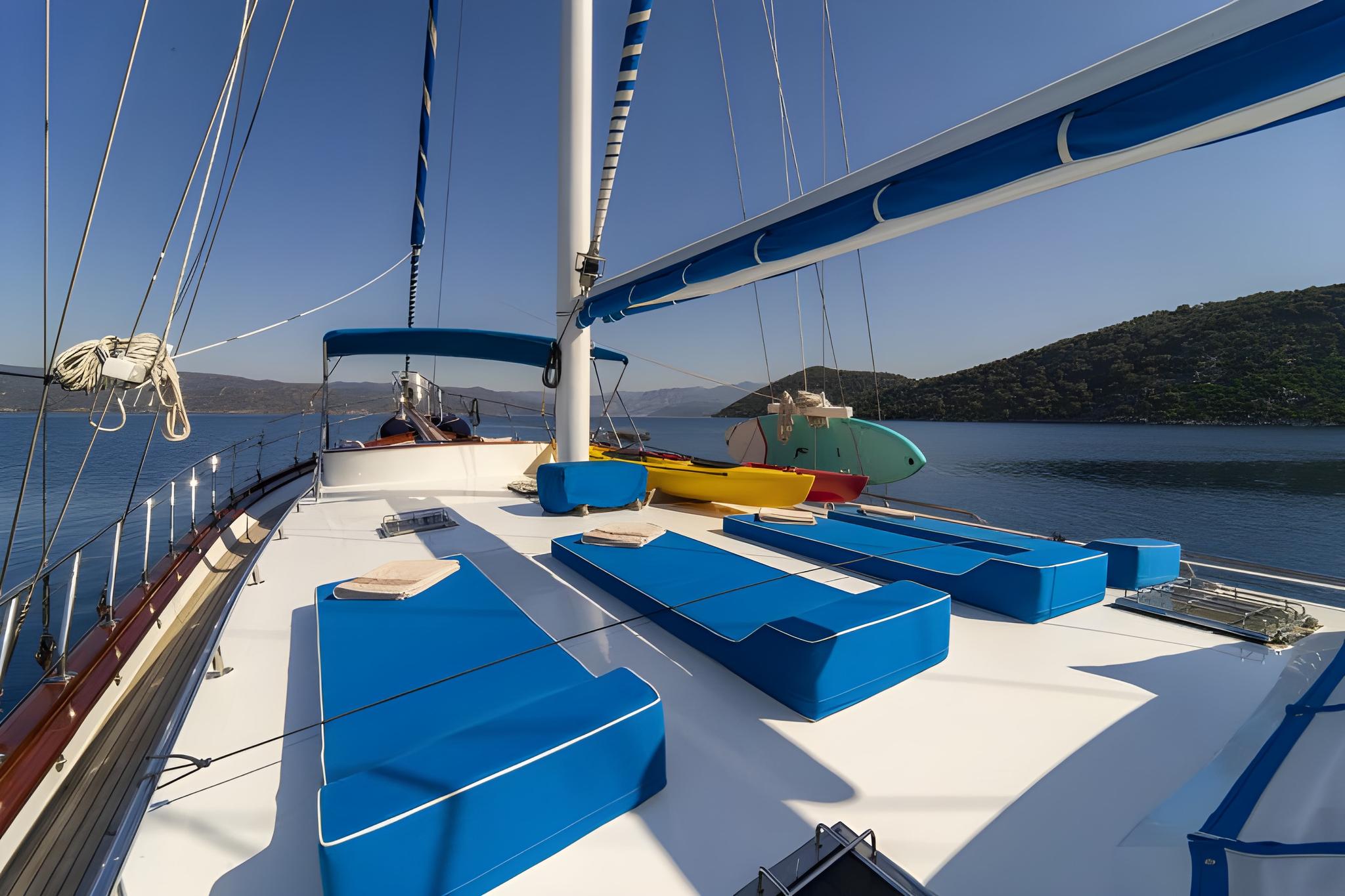 gulet bow deck with blue cushioned sun loungers and sailing rigging against coastal backdrop