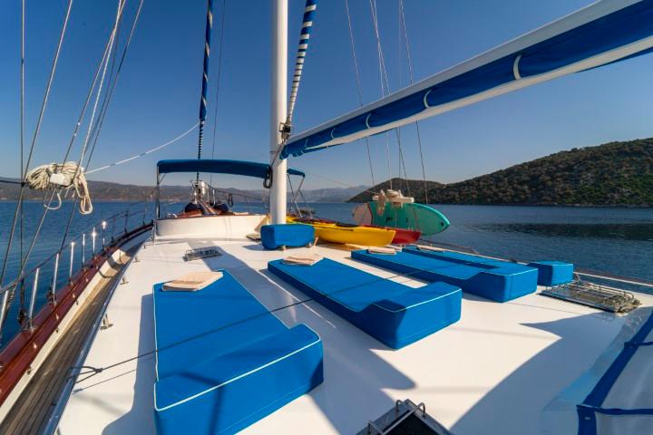 gulet bow deck with blue cushioned sun loungers and sailing rigging against coastal backdrop