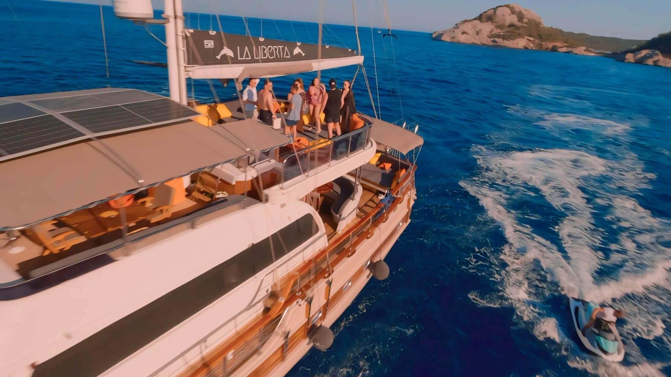 Aerial drone shot of gulet La Liberta underway with guests on sun deck and jet-ski in wake