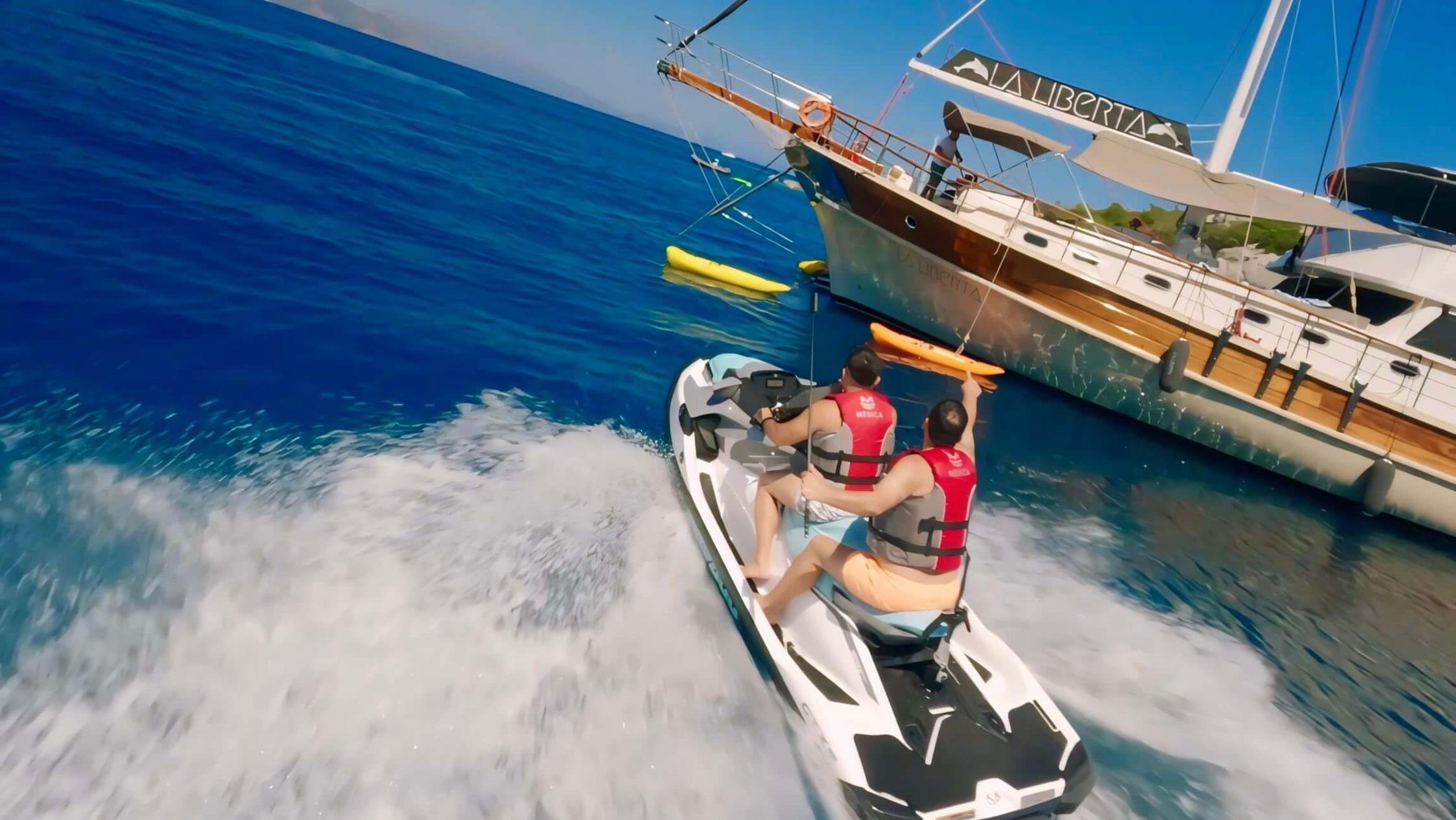 two guests in life jackets riding a jet ski alongside gulet La Liberta with yellow kayak visible